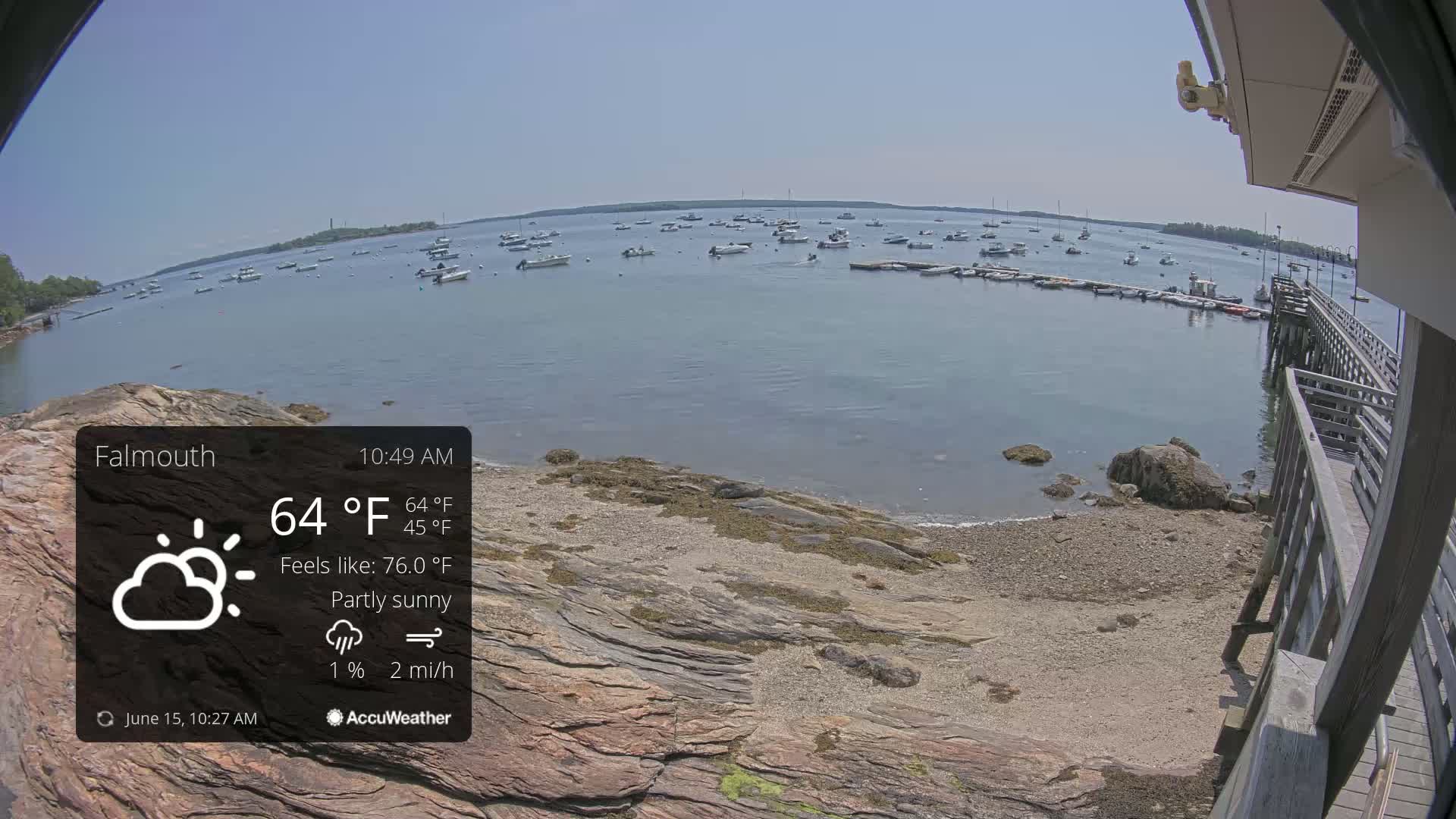 A partly sunny day with a gentle breeze shows a rocky shoreline with a calm body of water containing many small boats and a wooden pier.