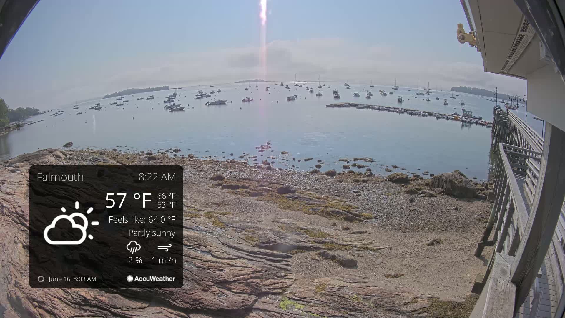 A partly sunny, calm day overlooks a rocky shoreline with numerous boats in a harbor.