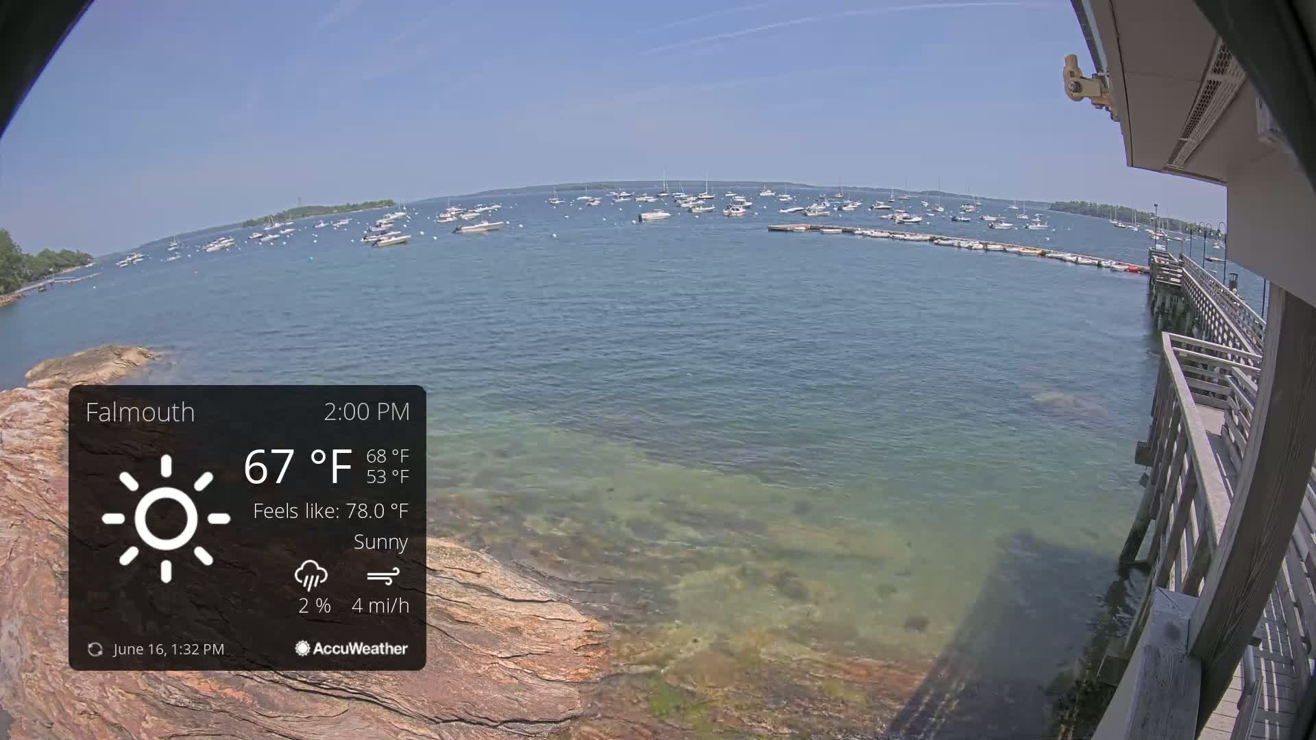 A sunny, calm day shows numerous boats at anchor in a bay, viewed from a wooden pier.