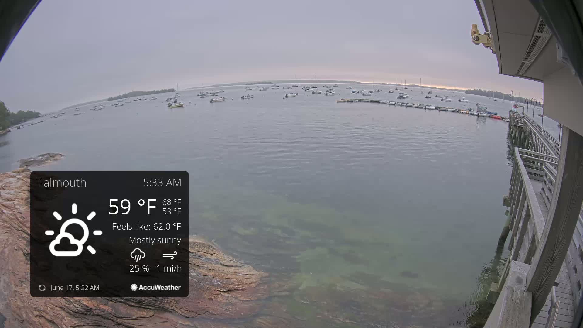A mostly sunny, calm morning scene shows a harbor filled with numerous boats, viewed from a wooden pier with clear, shallow water visible near the shore.