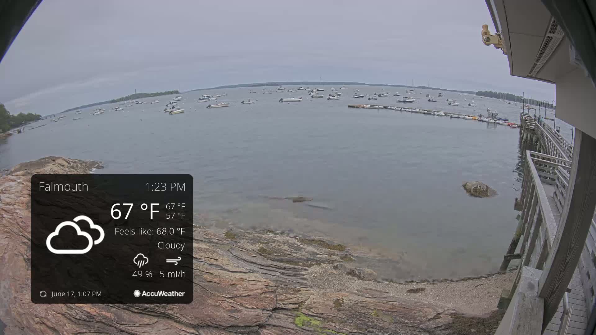 A cloudy, 67°F day shows a calm body of water with numerous boats moored, viewed from a rocky shoreline next to a wooden pier.
