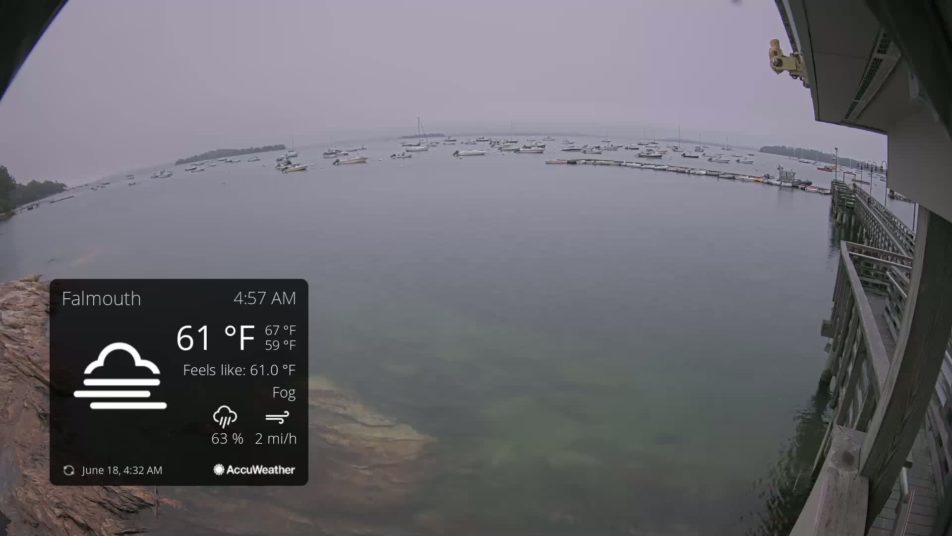 A foggy morning scene shows numerous boats moored in calm water, viewed from a wooden pier.