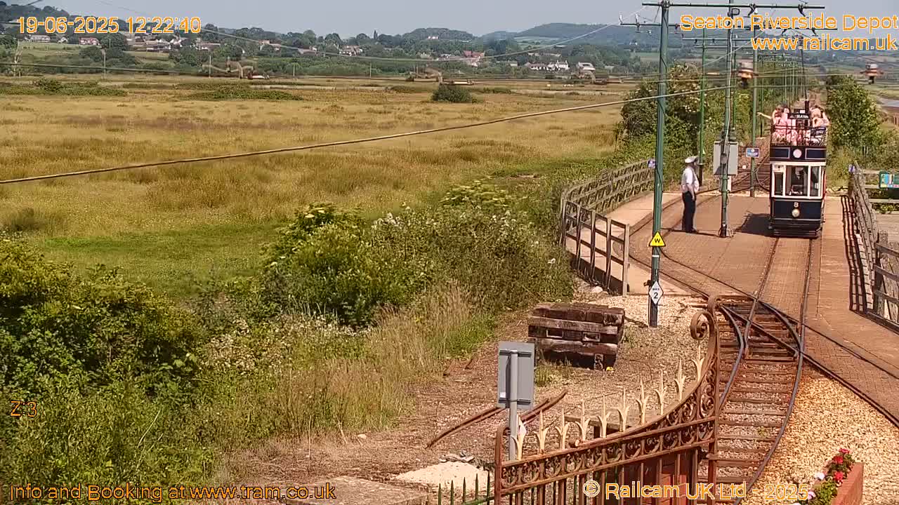 A tram carrying passengers approaches a station on a sunny day, passing alongside a grassy field and a small town in the distance.