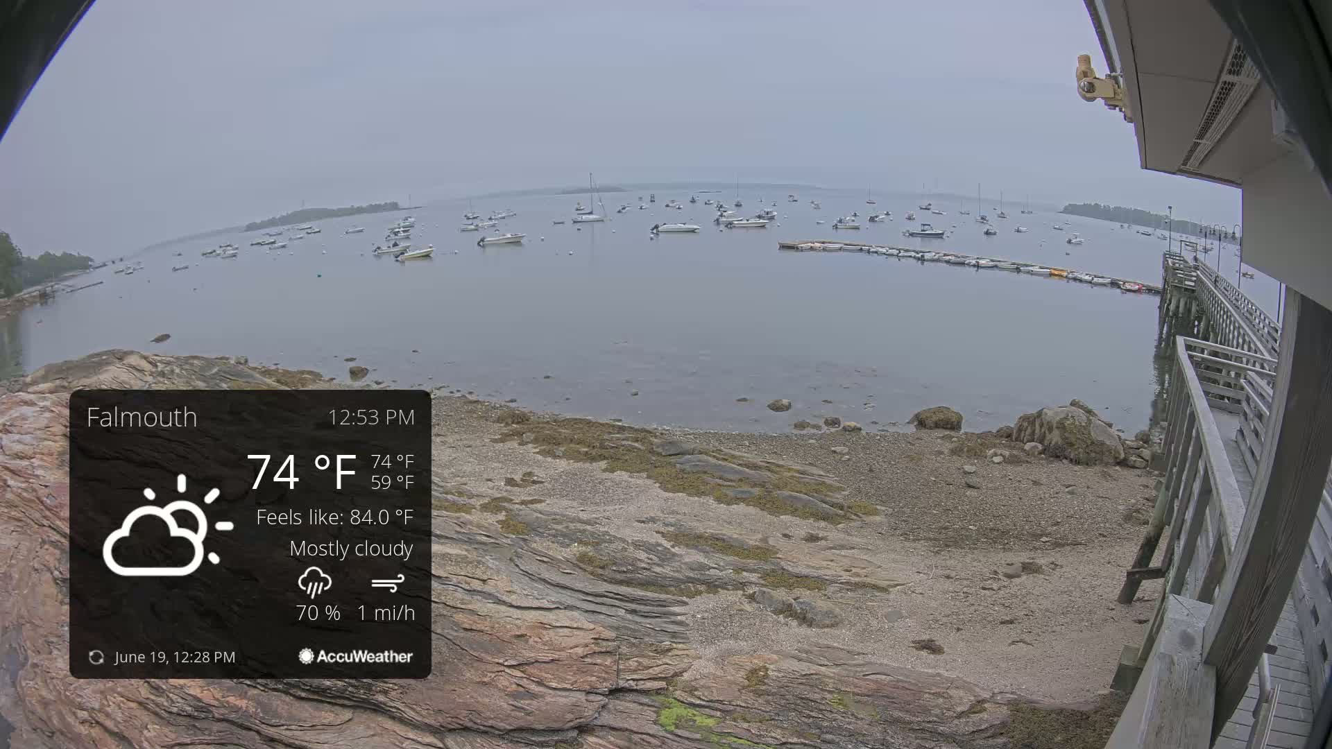 A mostly cloudy, 74°F day shows a calm bay with numerous boats moored near a rocky shoreline and a wooden pier.