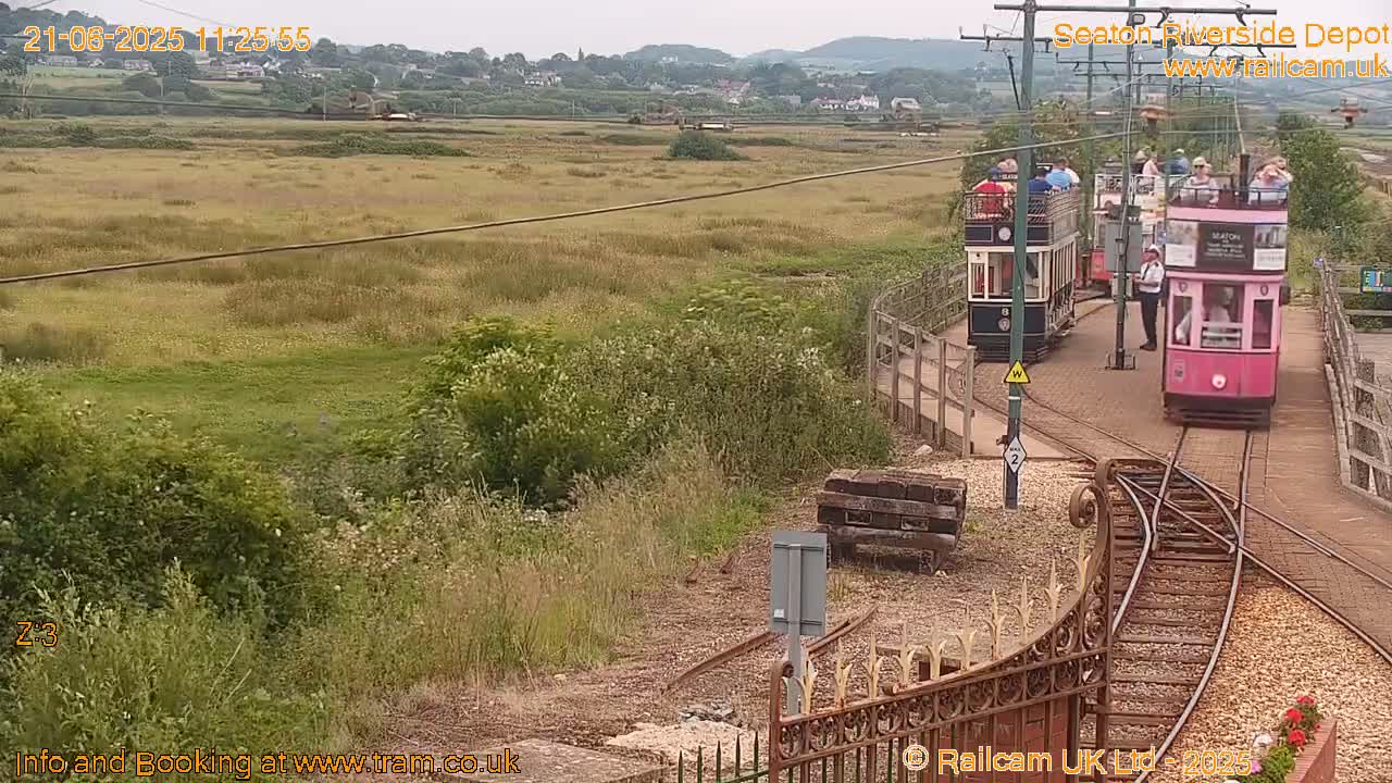Two passenger trams are traveling along a track on a partly sunny day, passing a grassy area with distant hills in the background.