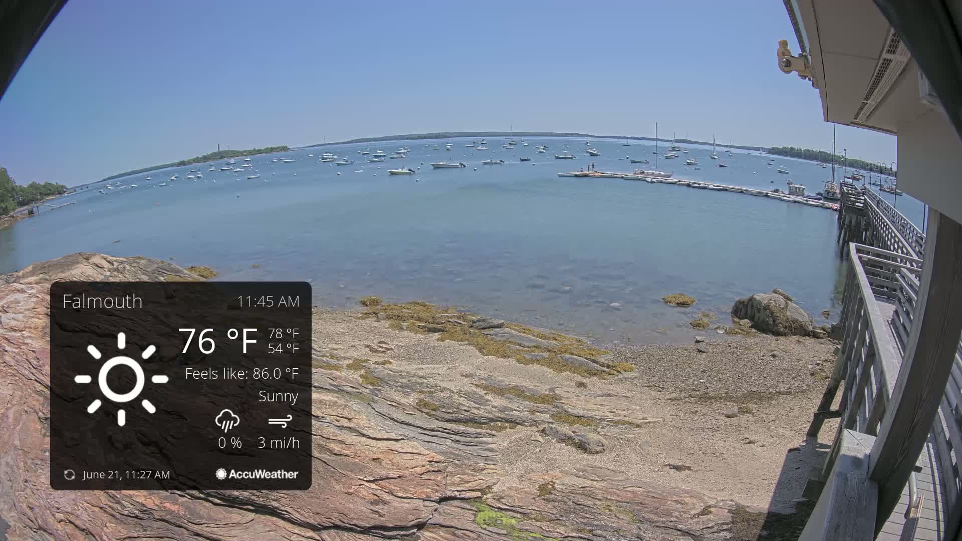 A sunny, calm bay with numerous boats is viewed from a rocky shoreline near a wooden pier.