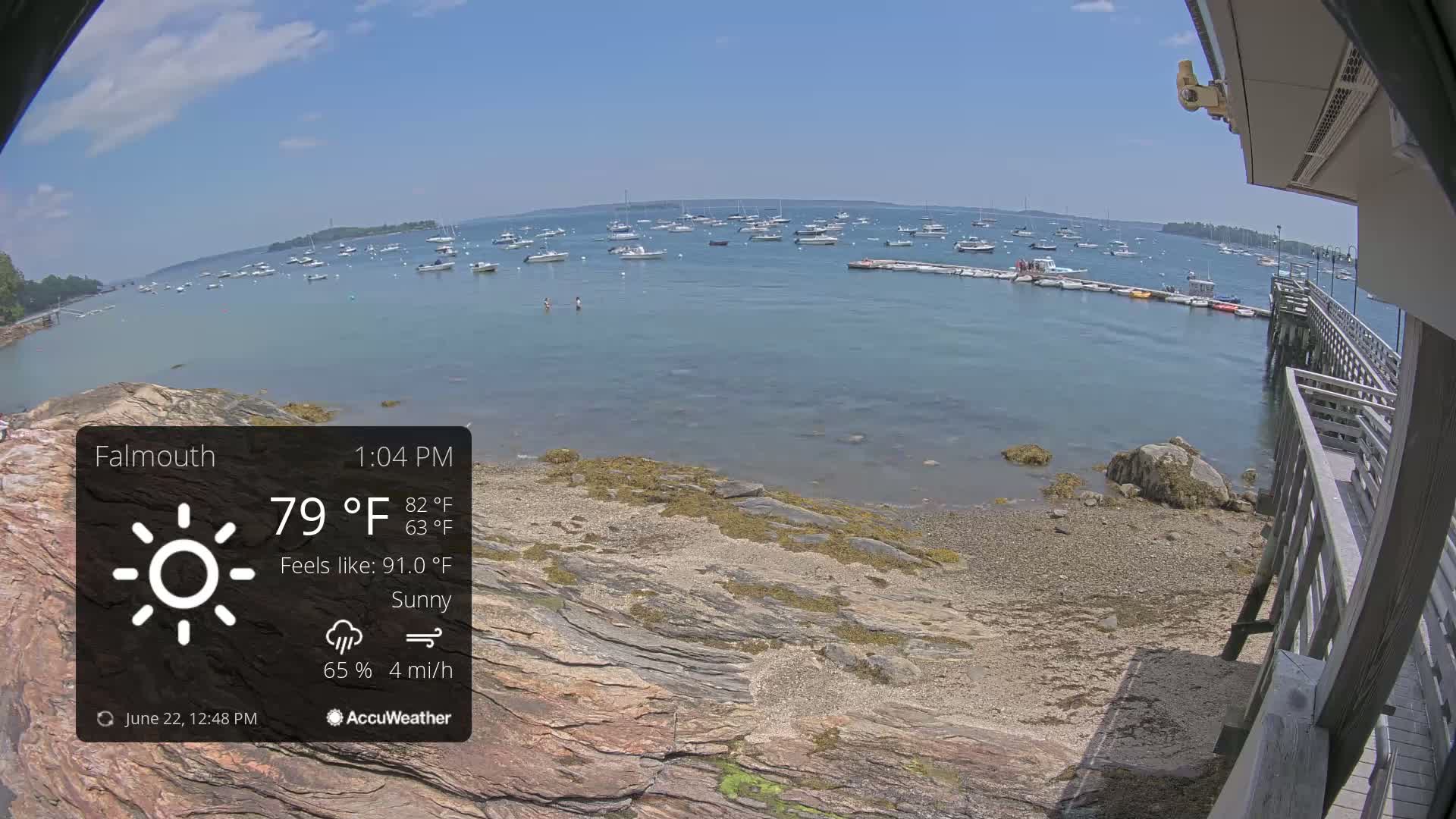 A sunny, breezy waterfront scene shows numerous boats in a calm bay, a rocky shoreline, and a wooden pier.