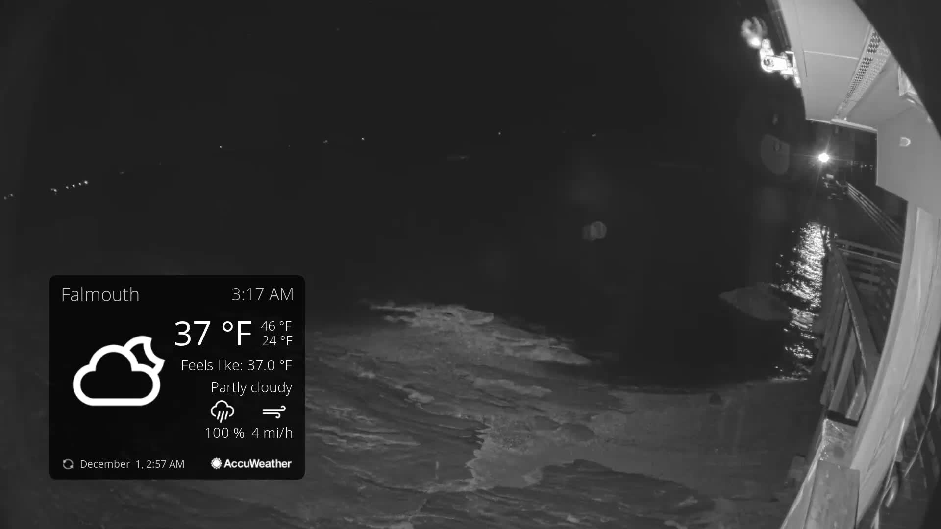 A dark, nighttime view shows choppy water next to a lit pier or vessel, with distant lights along the horizon under a partly cloudy sky.