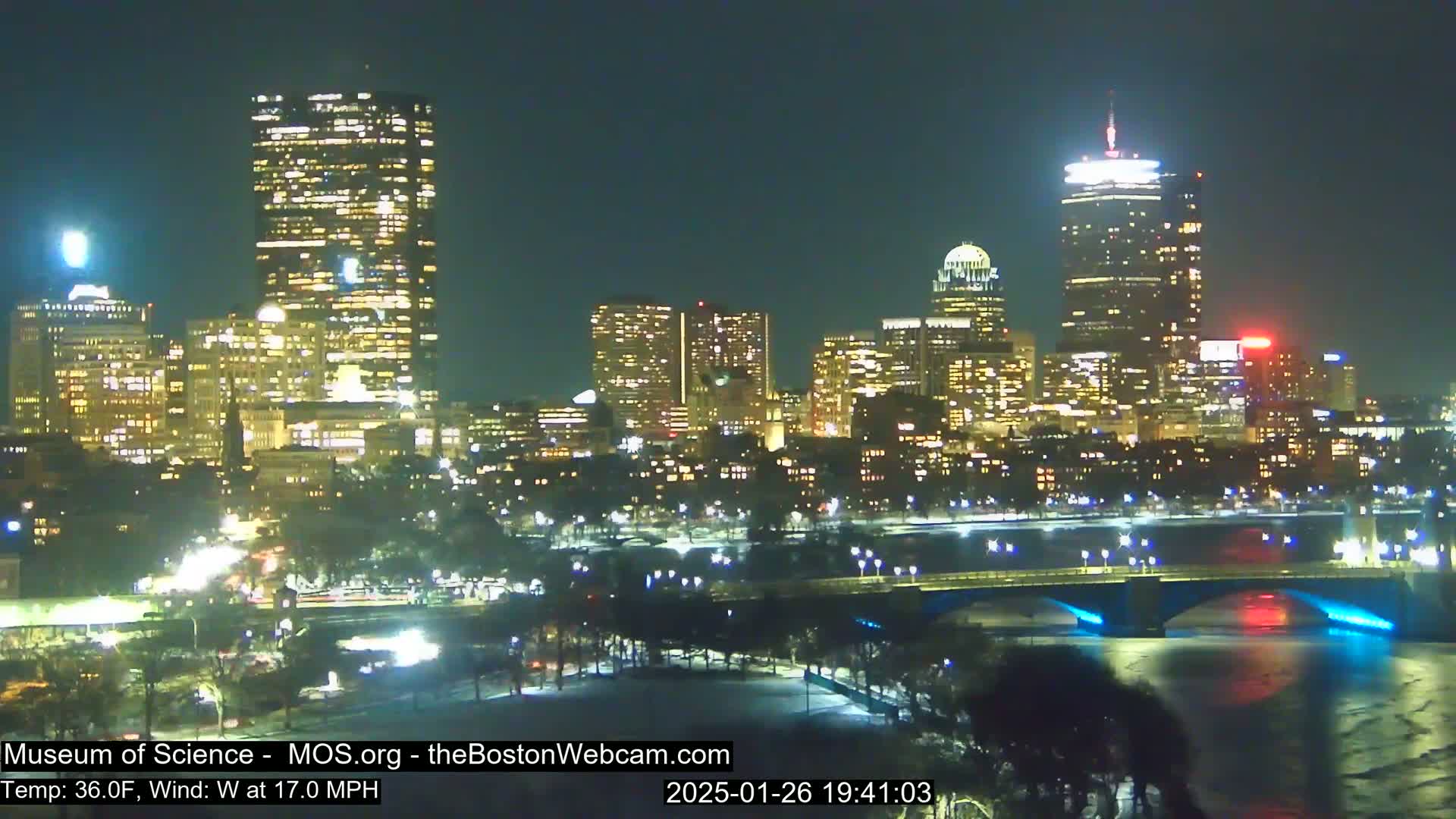 A nighttime view of a city skyline illuminated with lights, featuring a river and bridge, under a clear sky with a temperature of 36°F and a 17 mph west wind.