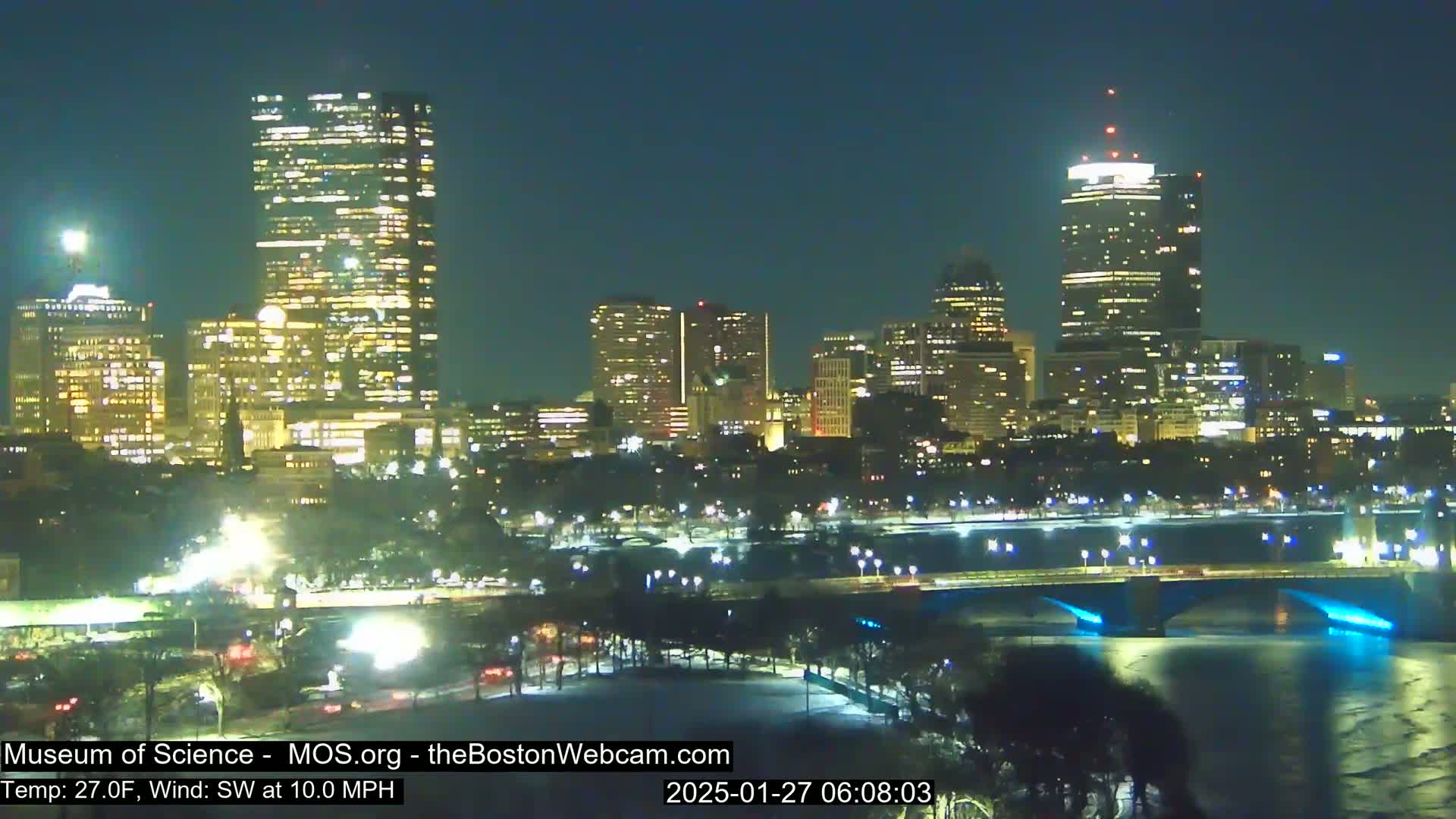 A nighttime view of a city skyline illuminated by lights, with a bridge over a body of water under a clear, dark sky and a temperature of 27°F, a 10 mph southwest wind.