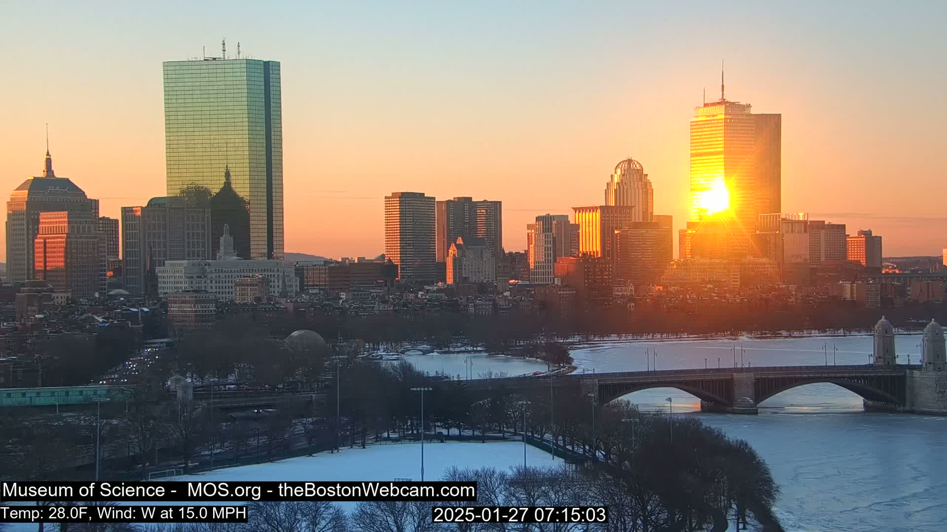 A city skyline with several tall buildings is visible at sunrise on a cold, windy day with a partially frozen river and a bridge in the foreground.