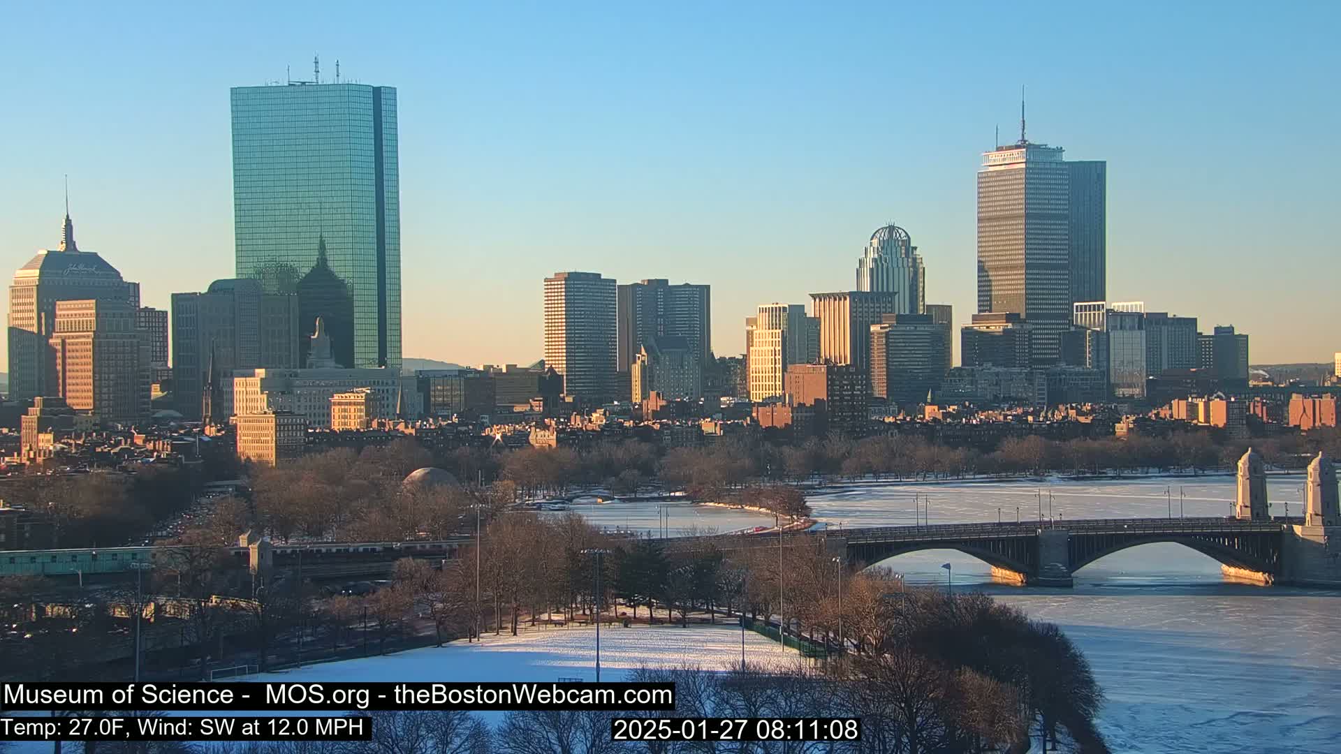 A city skyline with several skyscrapers is visible across a partially frozen river, under a clear, sunny sky.