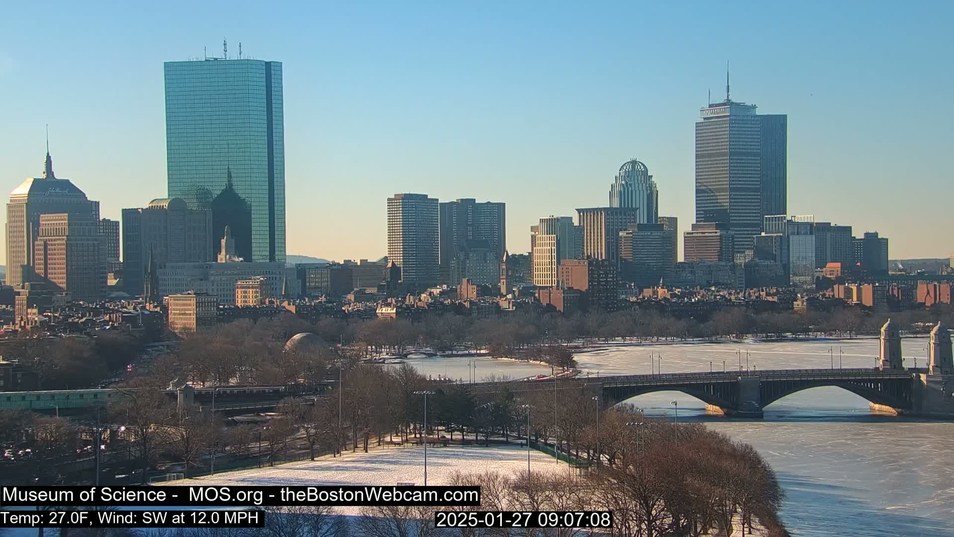 A snow-covered city skyline, featuring numerous skyscrapers and a bridge spanning a partially frozen river, is visible under a clear, sunny sky.