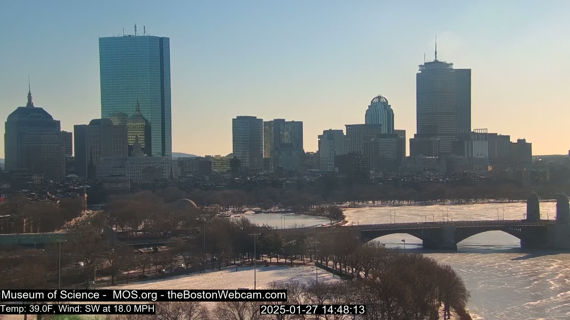 A city skyline with several tall buildings is visible across a partially frozen river, under a clear, sunny sky.