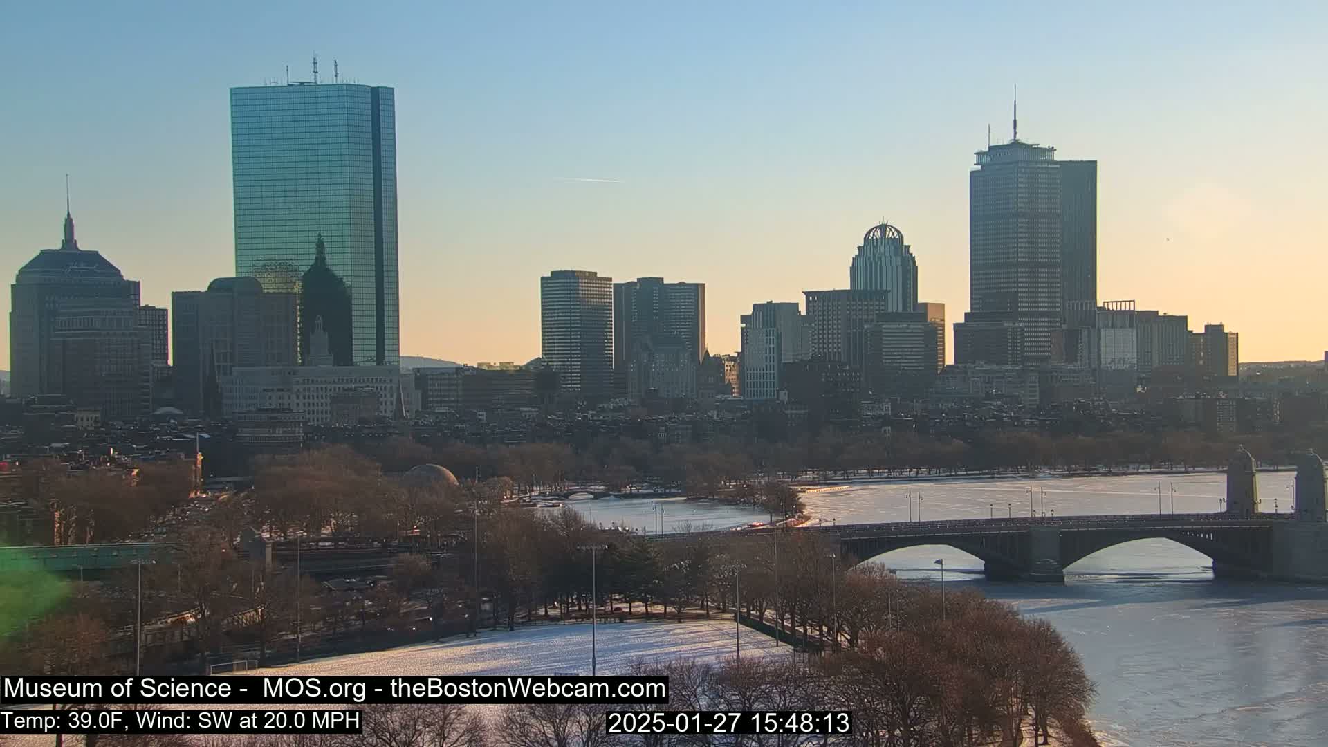 A cityscape with numerous skyscrapers is seen across a partially frozen river, with a bridge in the foreground, under a clear, light-blue sky.