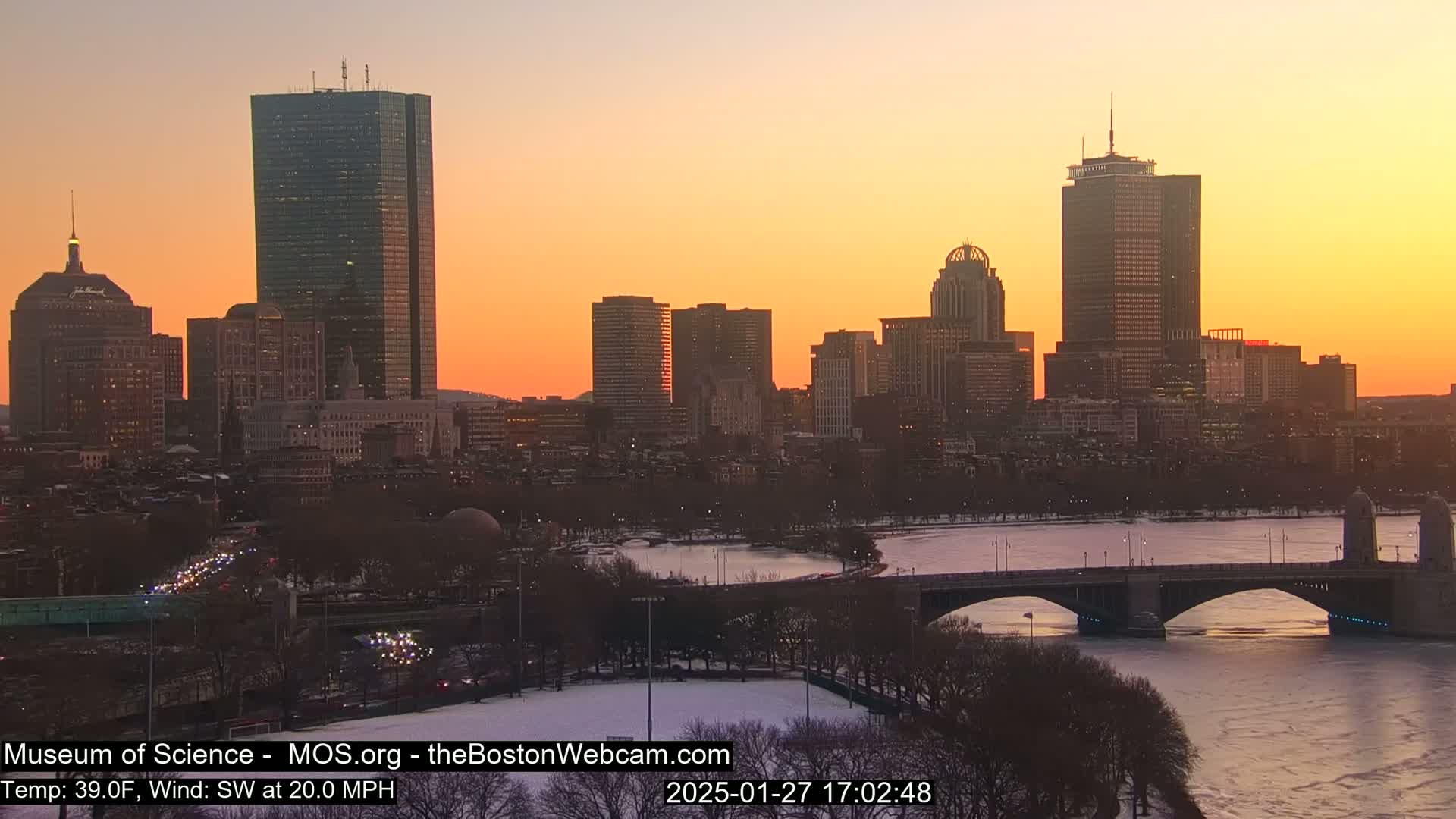 A city skyline at sunset, with a river partially frozen over and a bridge crossing it, under a clear, orange-tinged sky.
