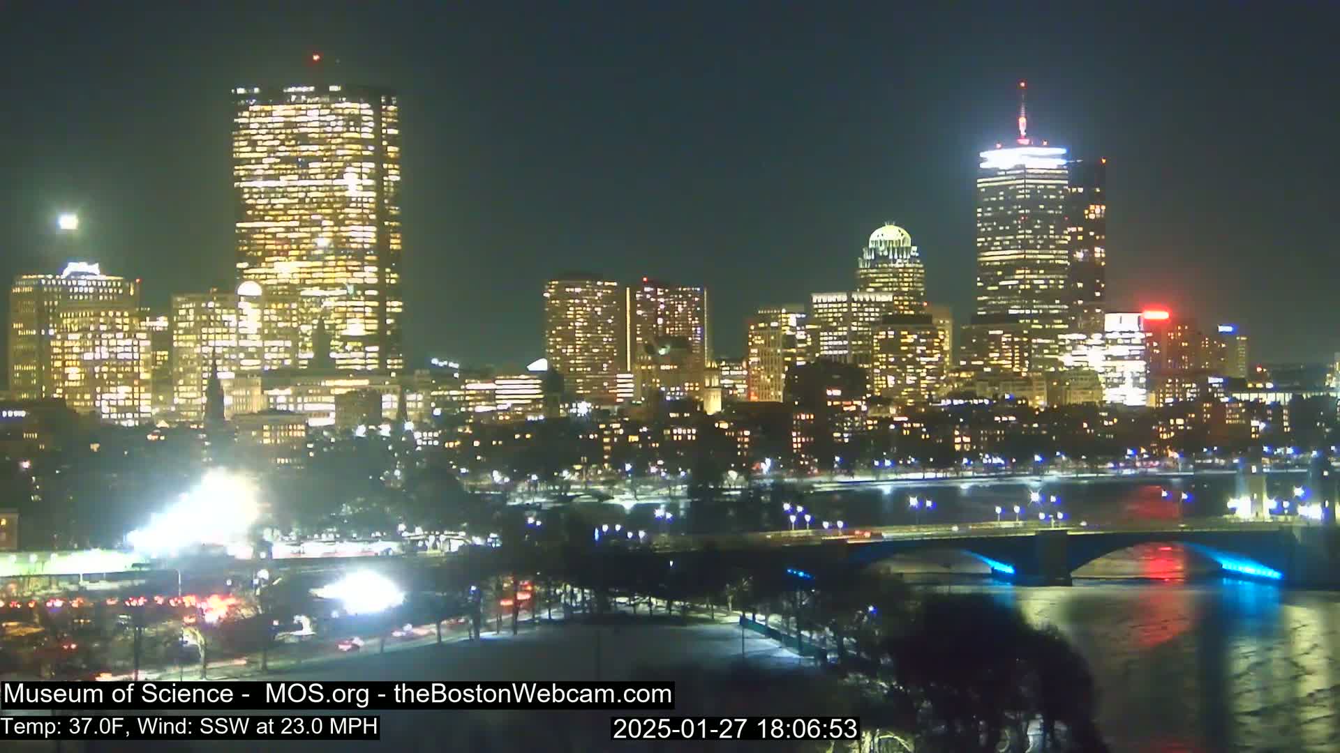 A nighttime view of a city skyline illuminated with lights, across a river with a bridge, under a clear sky with a 37°F temperature and a 23 mph south-southwest wind.