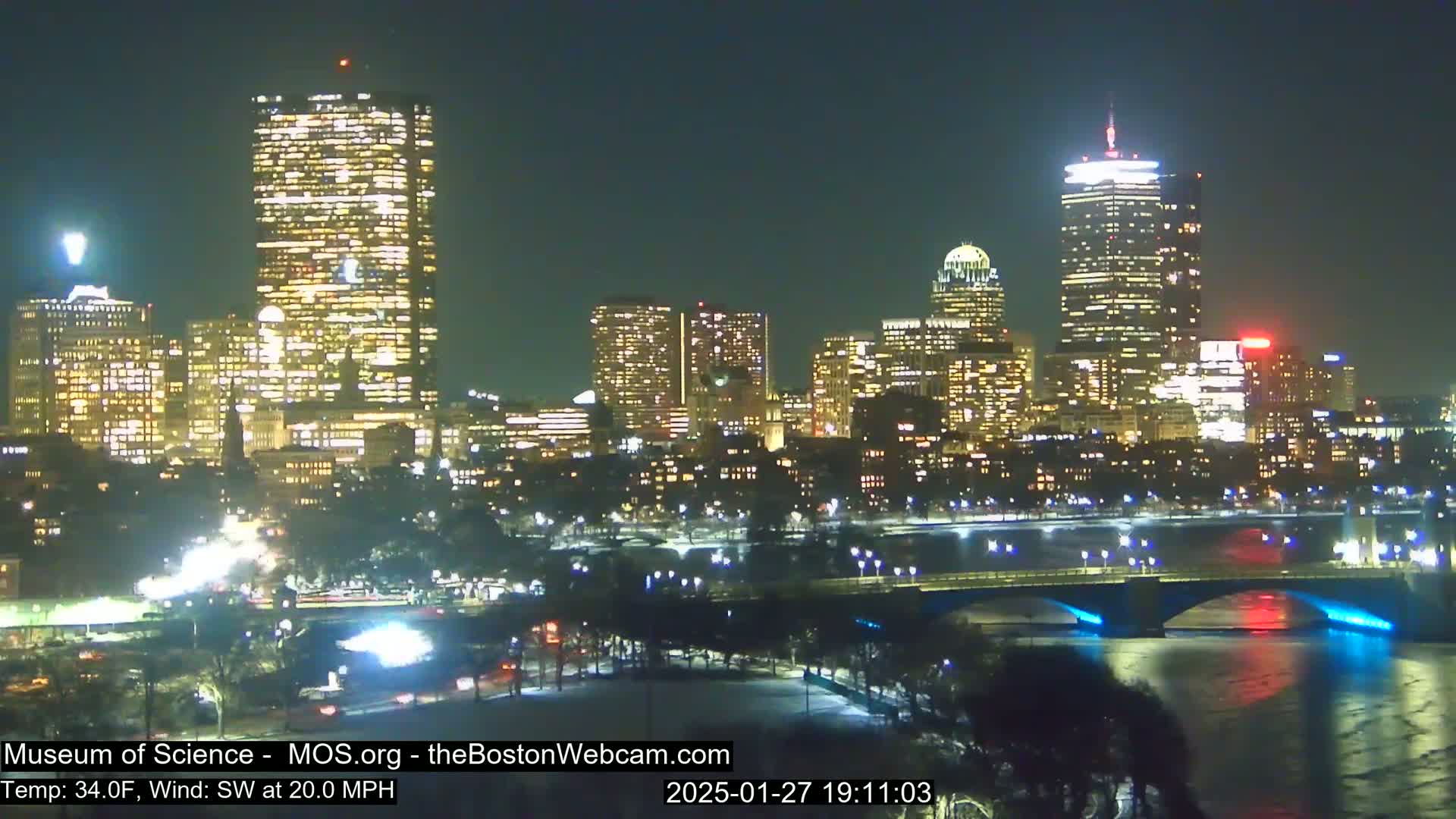 A nighttime view of a city skyline illuminated by lights, with a bridge spanning a dark river under a clear sky and a southwest wind at 20 mph.