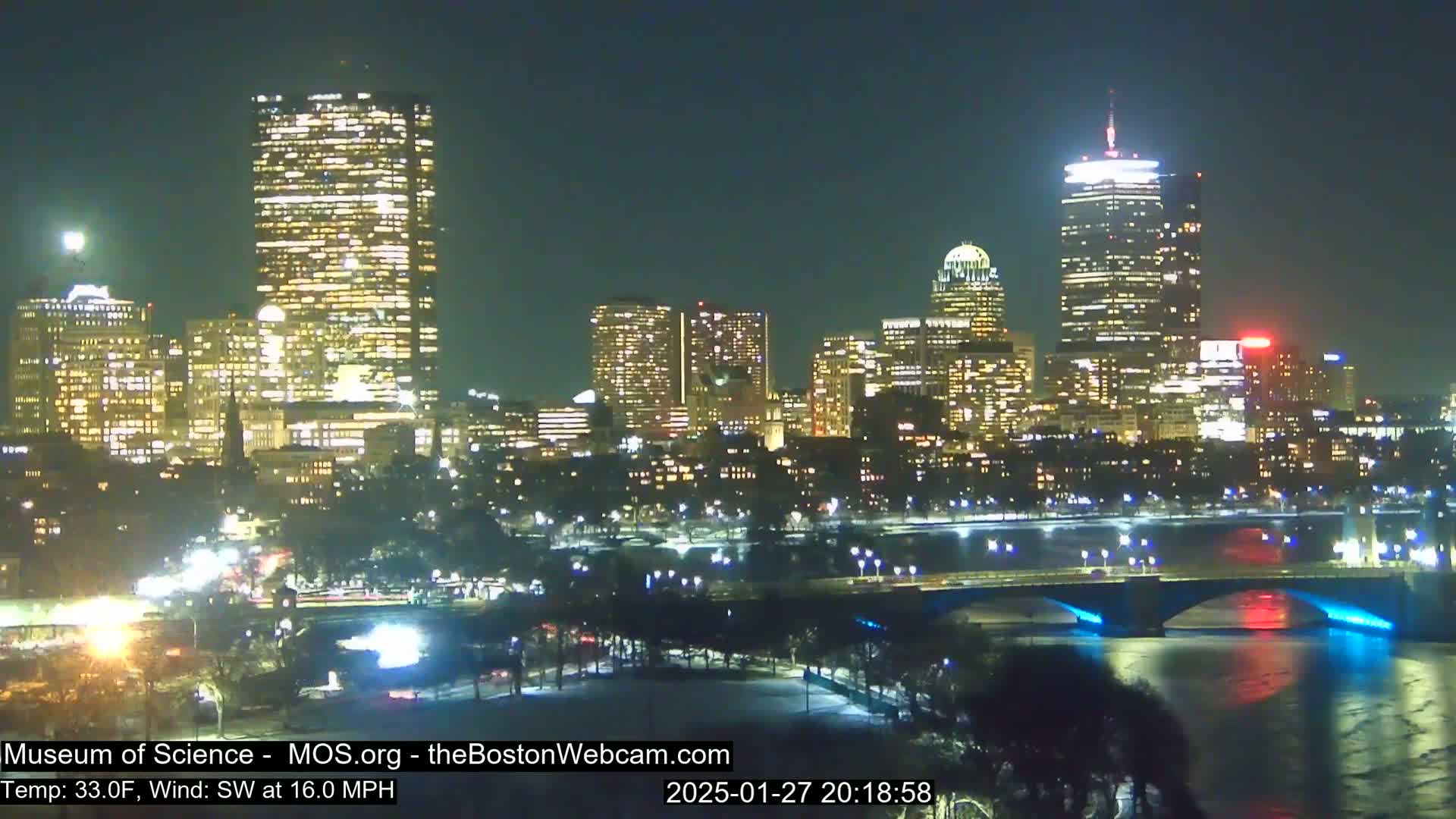 A nighttime view of a city skyline illuminated by lights, with a bridge spanning a body of water under a clear sky, and a temperature of 33 degrees Fahrenheit with a southwest wind of 16 mph.