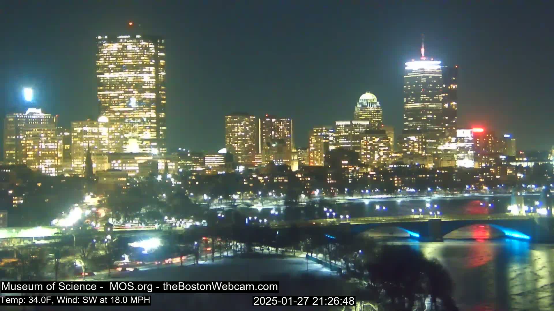 A nighttime cityscape with brightly lit buildings, a bridge, and a river under a clear sky, with a temperature of 34°F and a southwest wind at 18 mph.