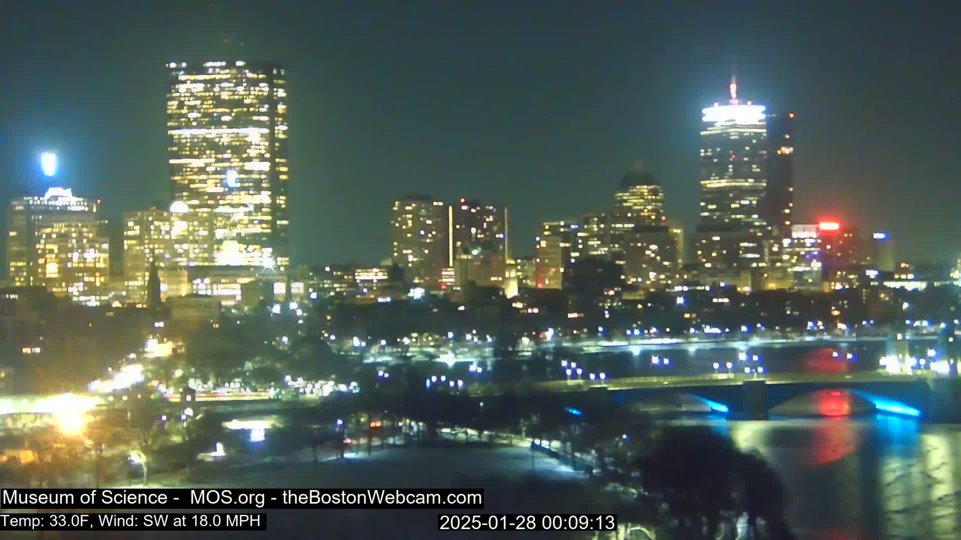 A nighttime cityscape with brightly lit skyscrapers and a bridge over a body of water, under a clear sky with a southwest wind of 18 mph and a temperature of 33°F.