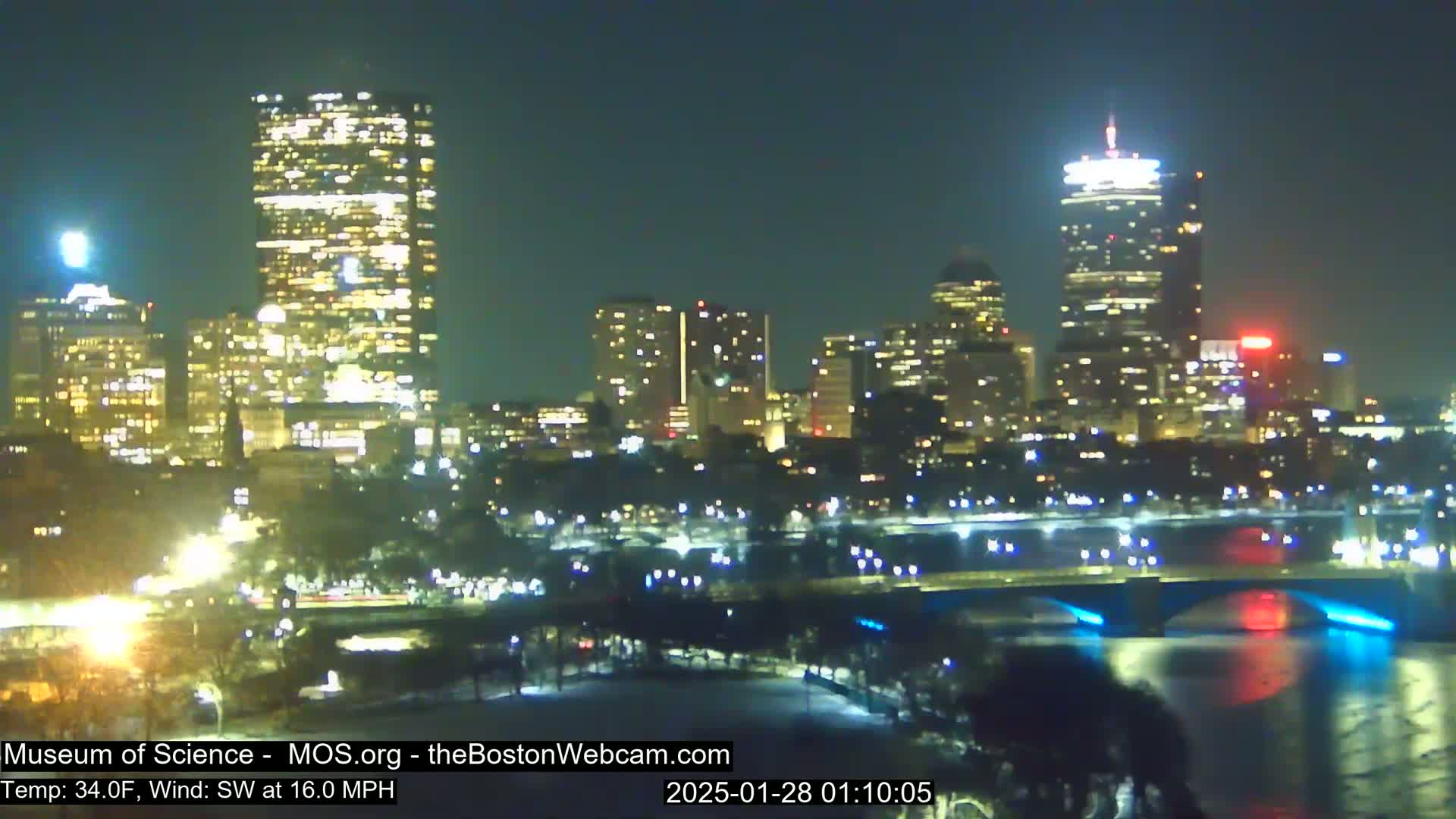 A nighttime cityscape with brightly lit buildings, a bridge, and a body of water under a clear sky, with a southwest wind blowing at 16 mph and a temperature of 34°F.