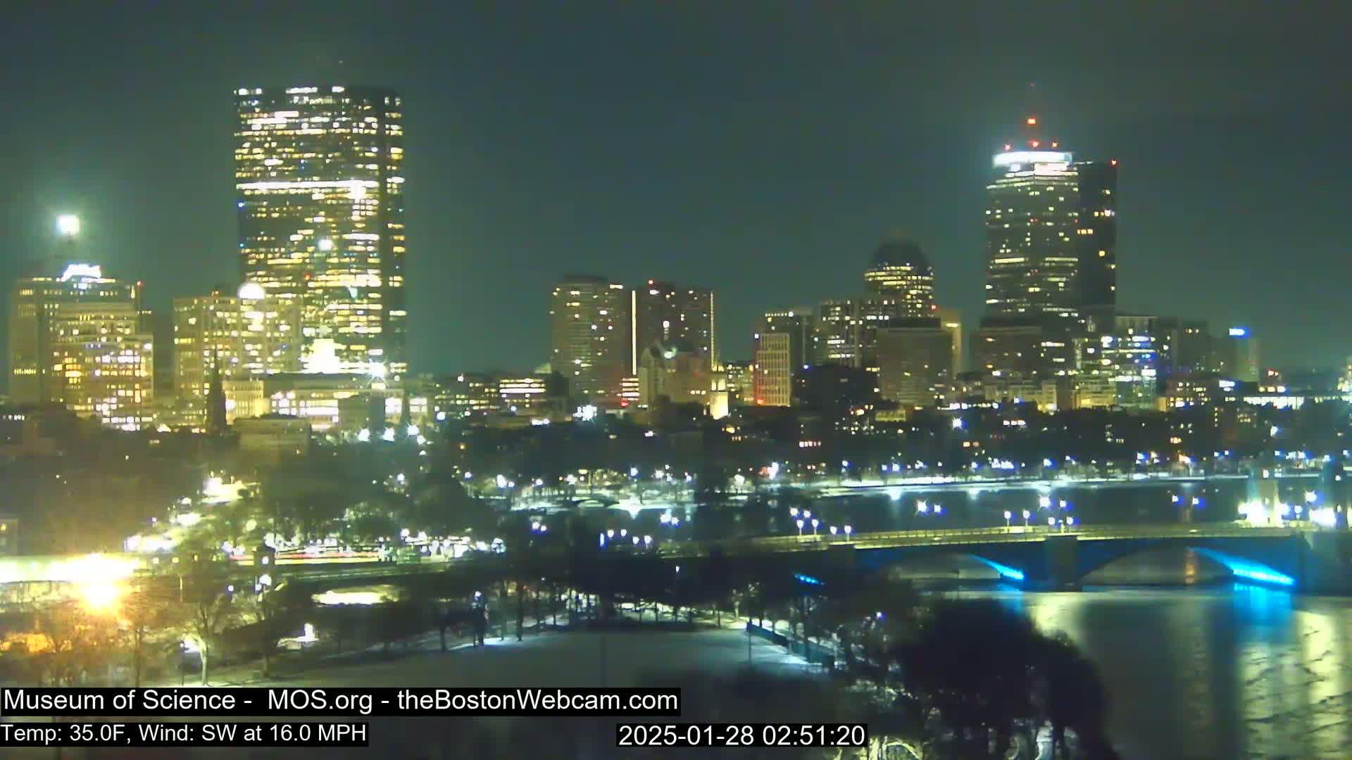 A nighttime cityscape with illuminated skyscrapers and bridges over a river, under a clear sky with a 35°F temperature and a 16 mph southwest wind.