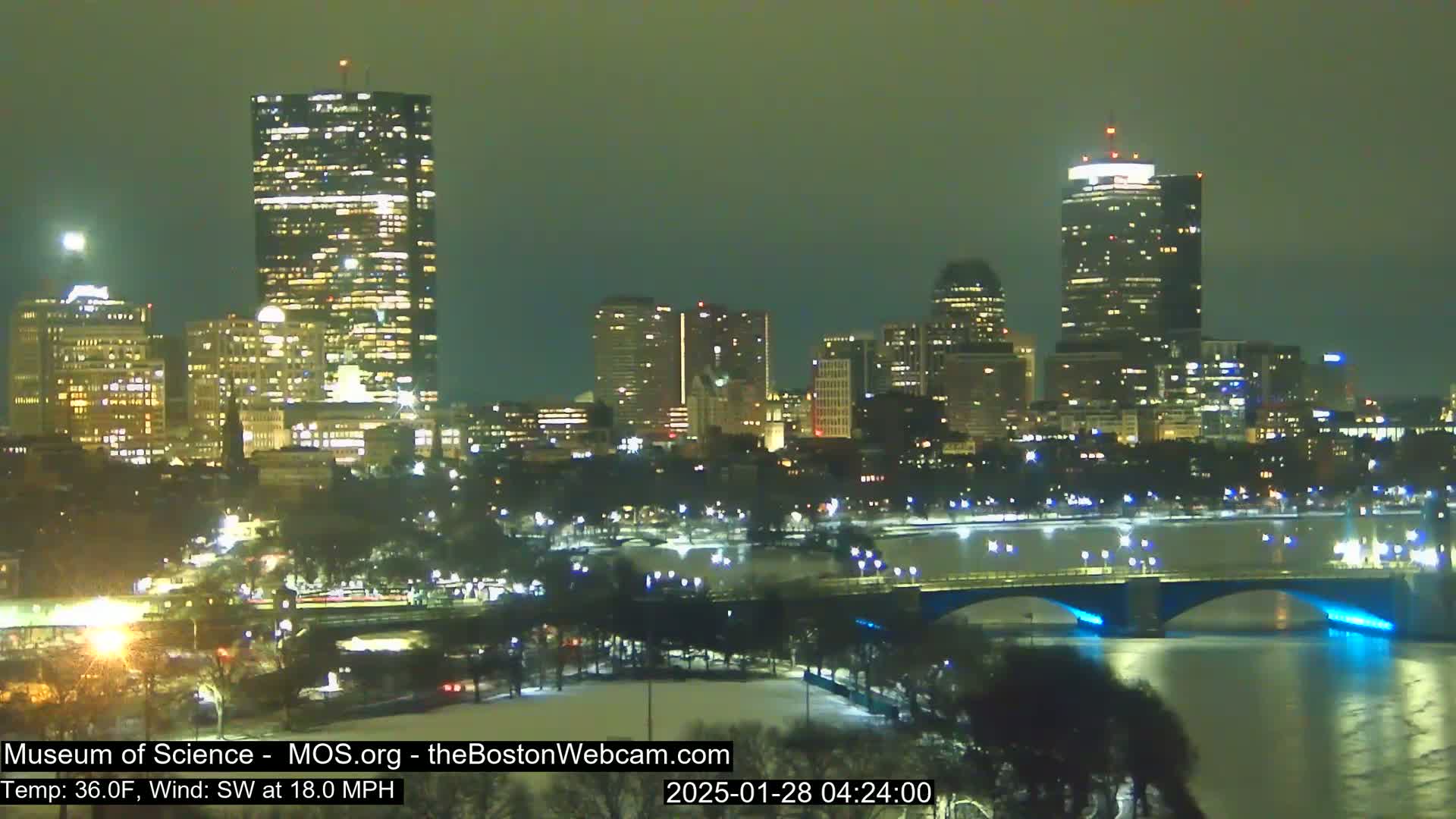 A nighttime view of a city skyline across a river with a bridge, under a dark, clear sky with a southwest wind at 18 mph and a temperature of 36 degrees Fahrenheit.