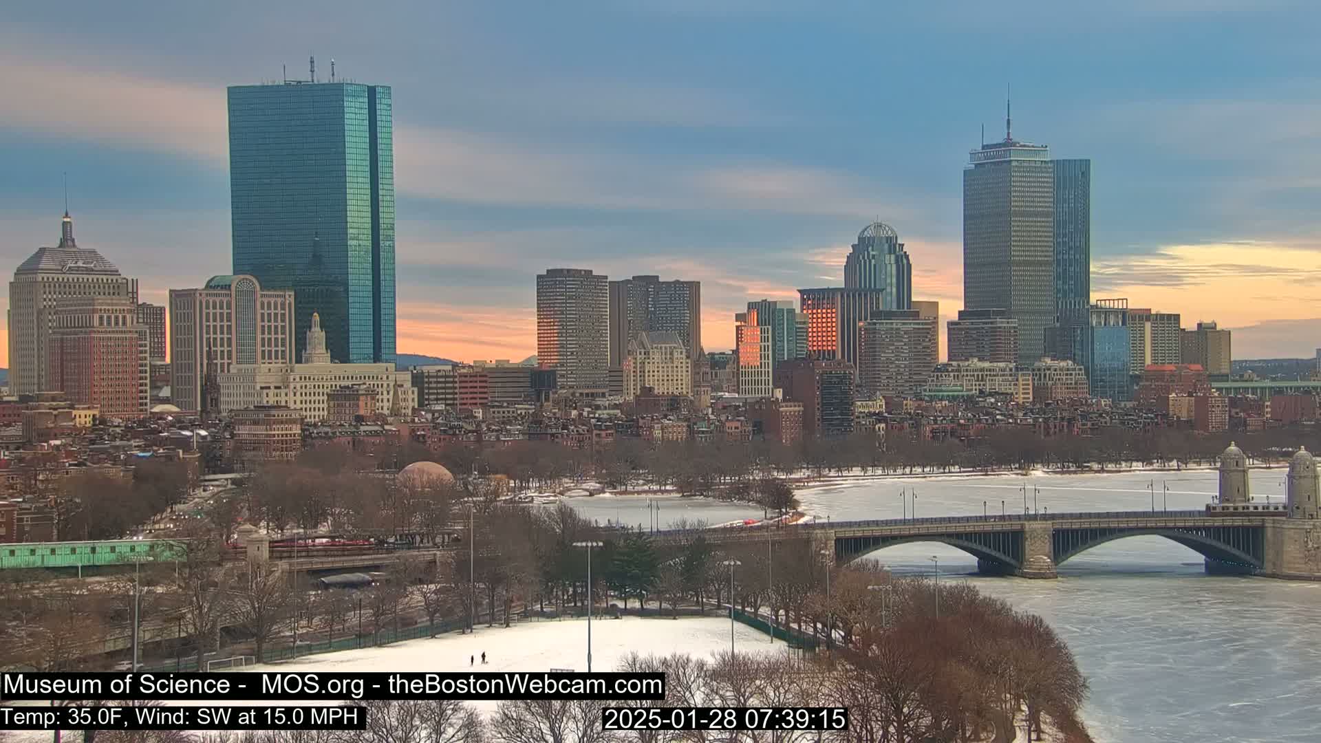 A snow-covered cityscape at sunrise, with a river partially frozen over and a bridge crossing it, under a partly cloudy sky.