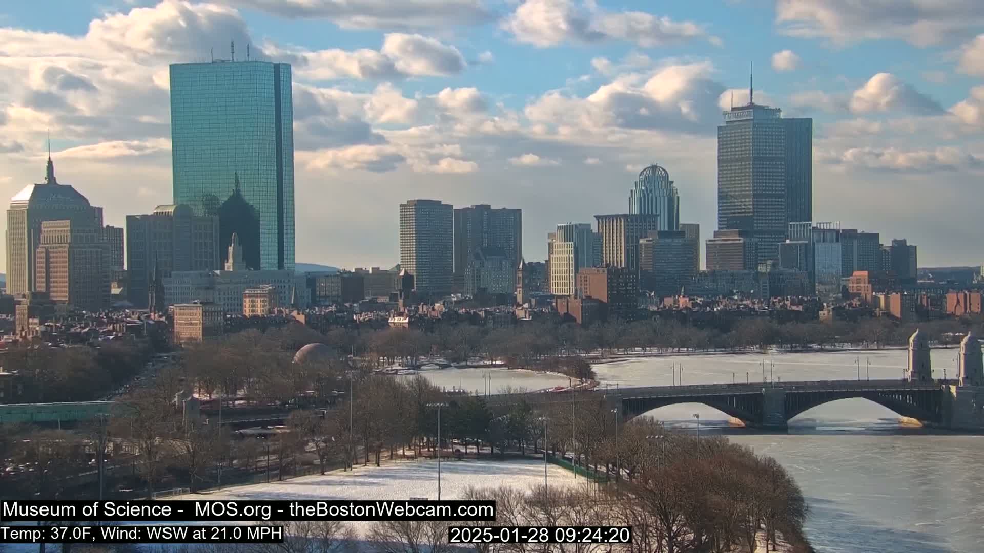 A partly cloudy day with a 37°F temperature and a 21 mph wind shows a city skyline with a bridge spanning a partially frozen river.