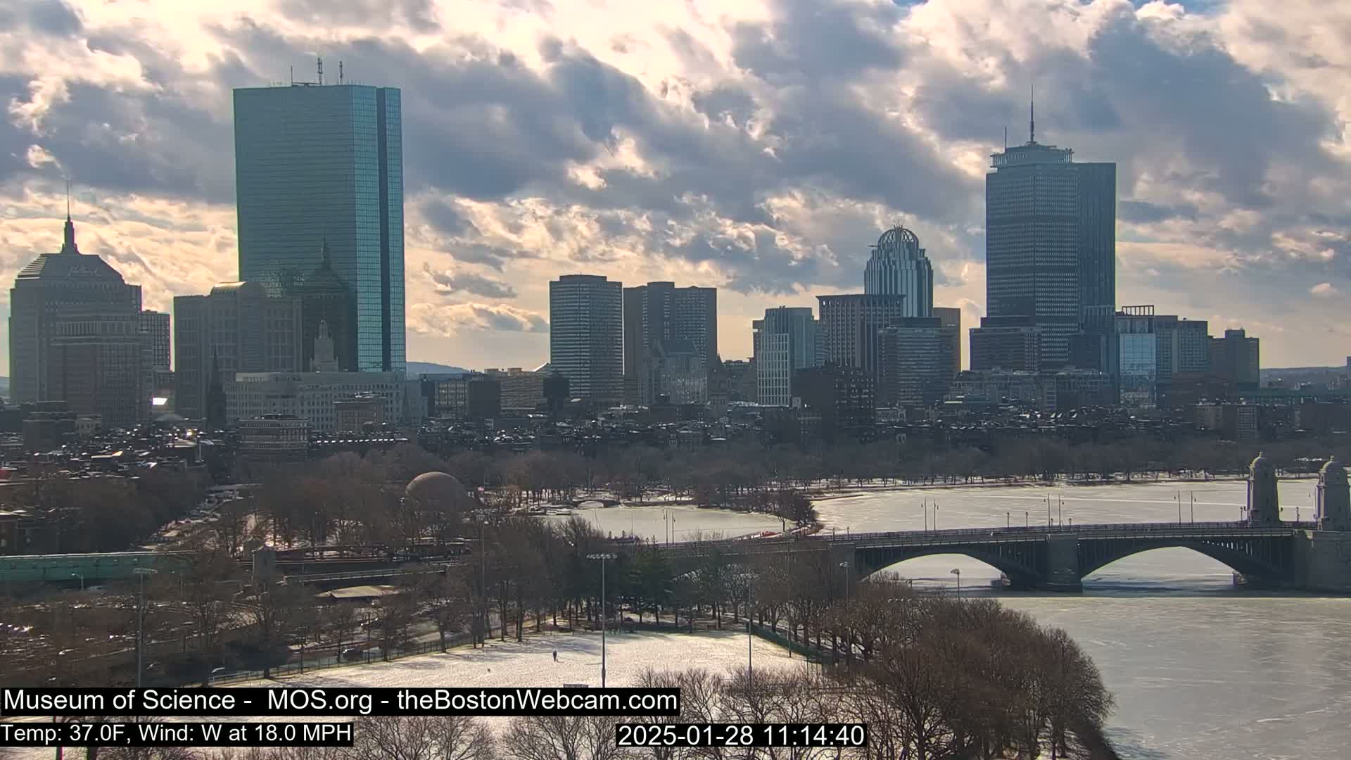A partly sunny day with an 18 mph west wind shows a city skyline across a partially frozen river with a bridge.