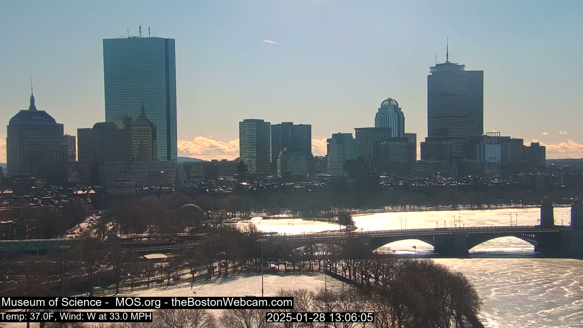 A sunny day with a 33 mph westerly wind shows a snow-covered park, a partly frozen river, and a bridge leading to a cityscape of tall buildings under a mostly clear sky.