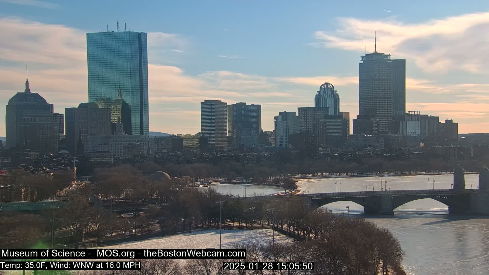 A partly sunny winter day shows the Boston skyline across a partially frozen river with a bridge in the foreground.