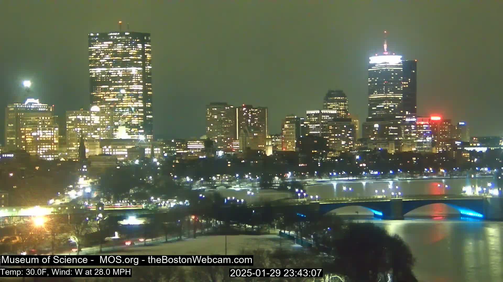 A nighttime cityscape with illuminated skyscrapers and a bridge over a river, under a dark, possibly overcast sky with a 30°F temperature and 28 mph west wind.