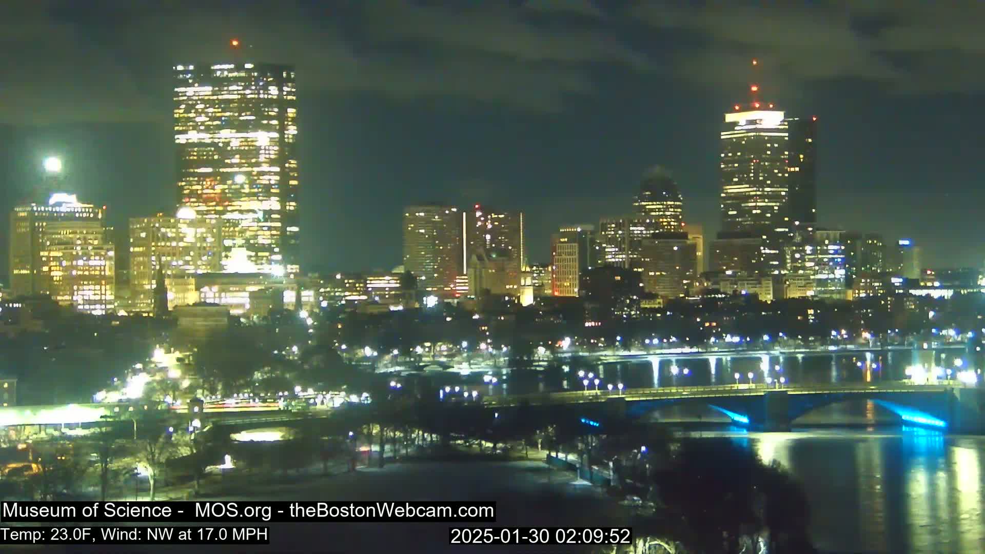 A nighttime view of a city skyline across a river, with a bridge illuminated in blue, under a partly cloudy sky.