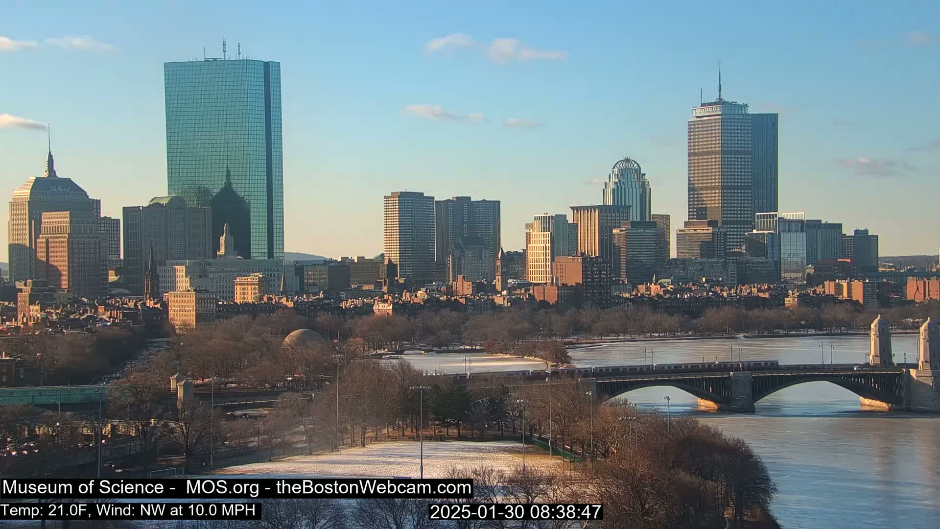 A sunny winter cityscape with a partially frozen river and a bridge carrying a train, showcasing numerous skyscrapers and buildings.