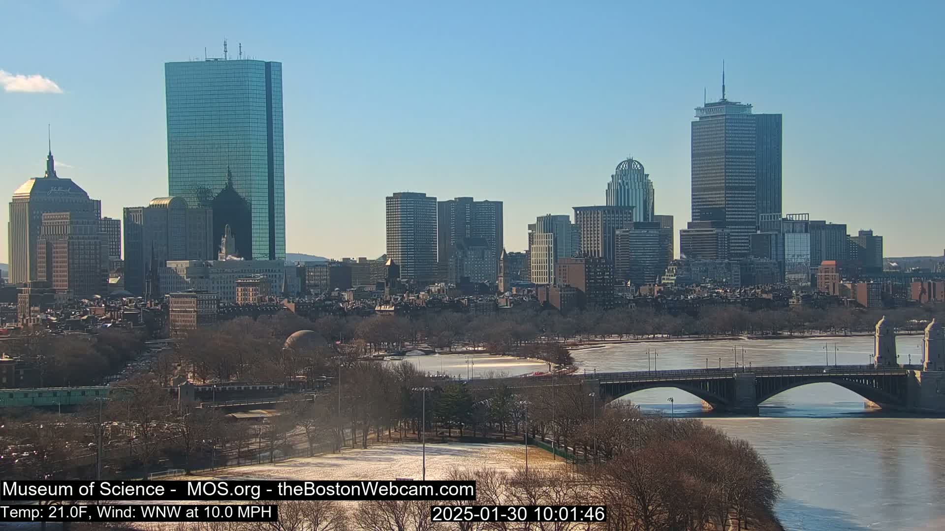 A sunny cityscape with a partially frozen river and bridge, under a clear blue sky.