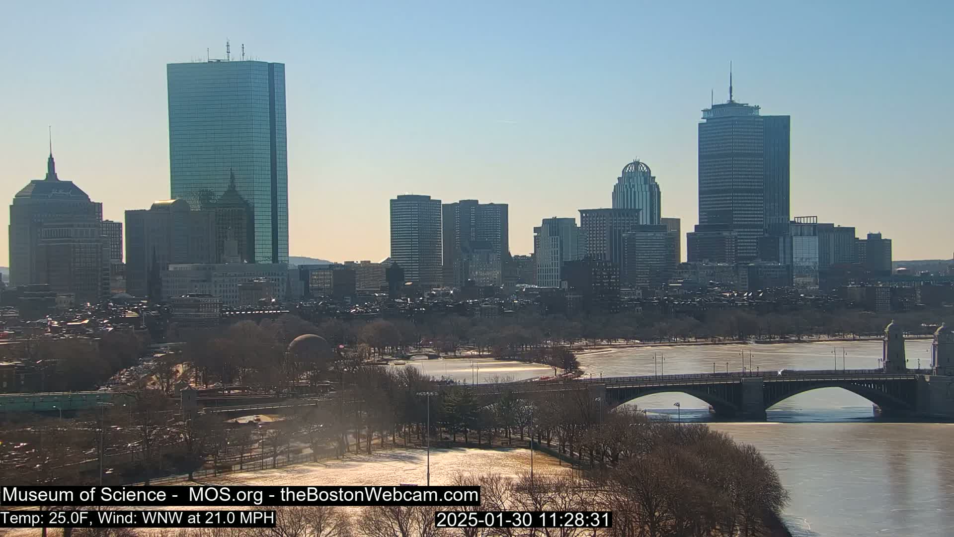 A sunny cityscape with a partly icy river and a bridge is seen under a clear blue sky, with a strong wind.