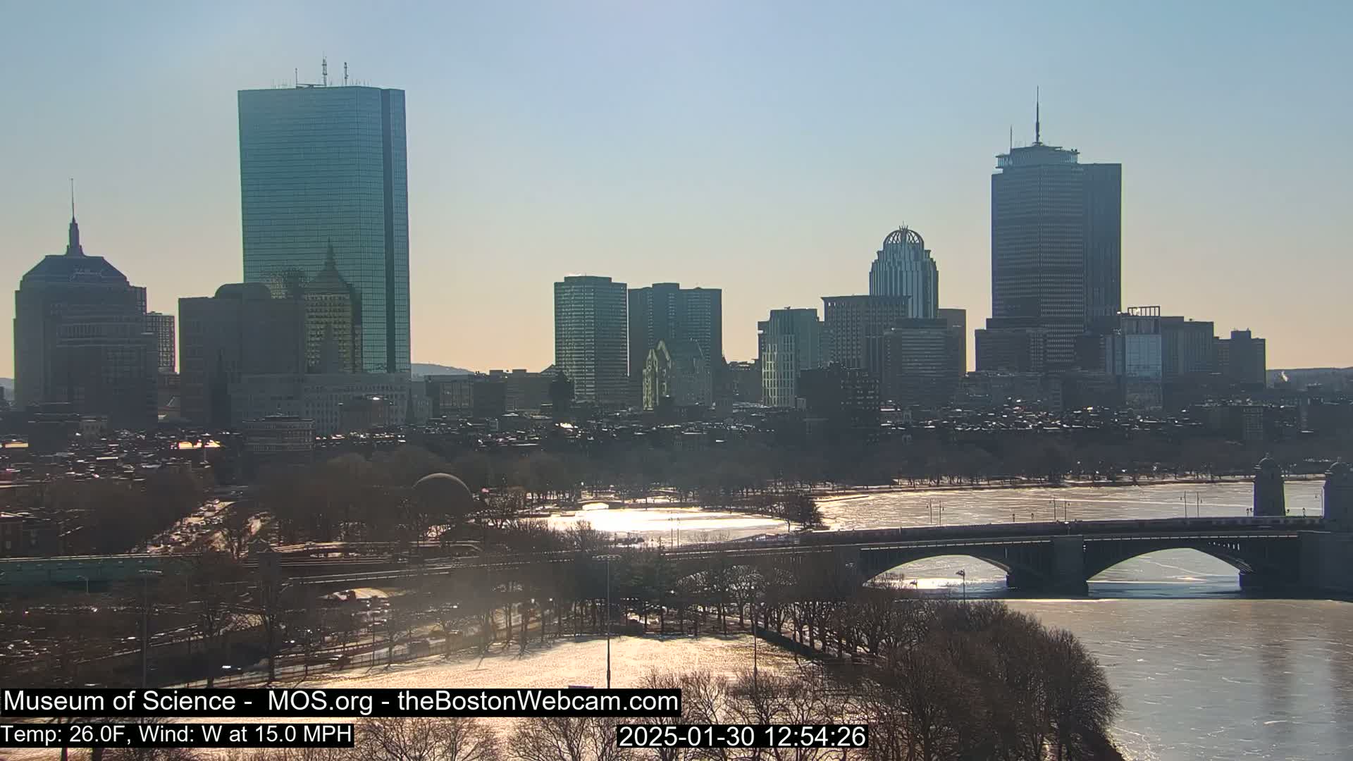 A sunny, cold, and windy day reveals a cityscape with numerous skyscrapers and a partially frozen river, crossed by a bridge.