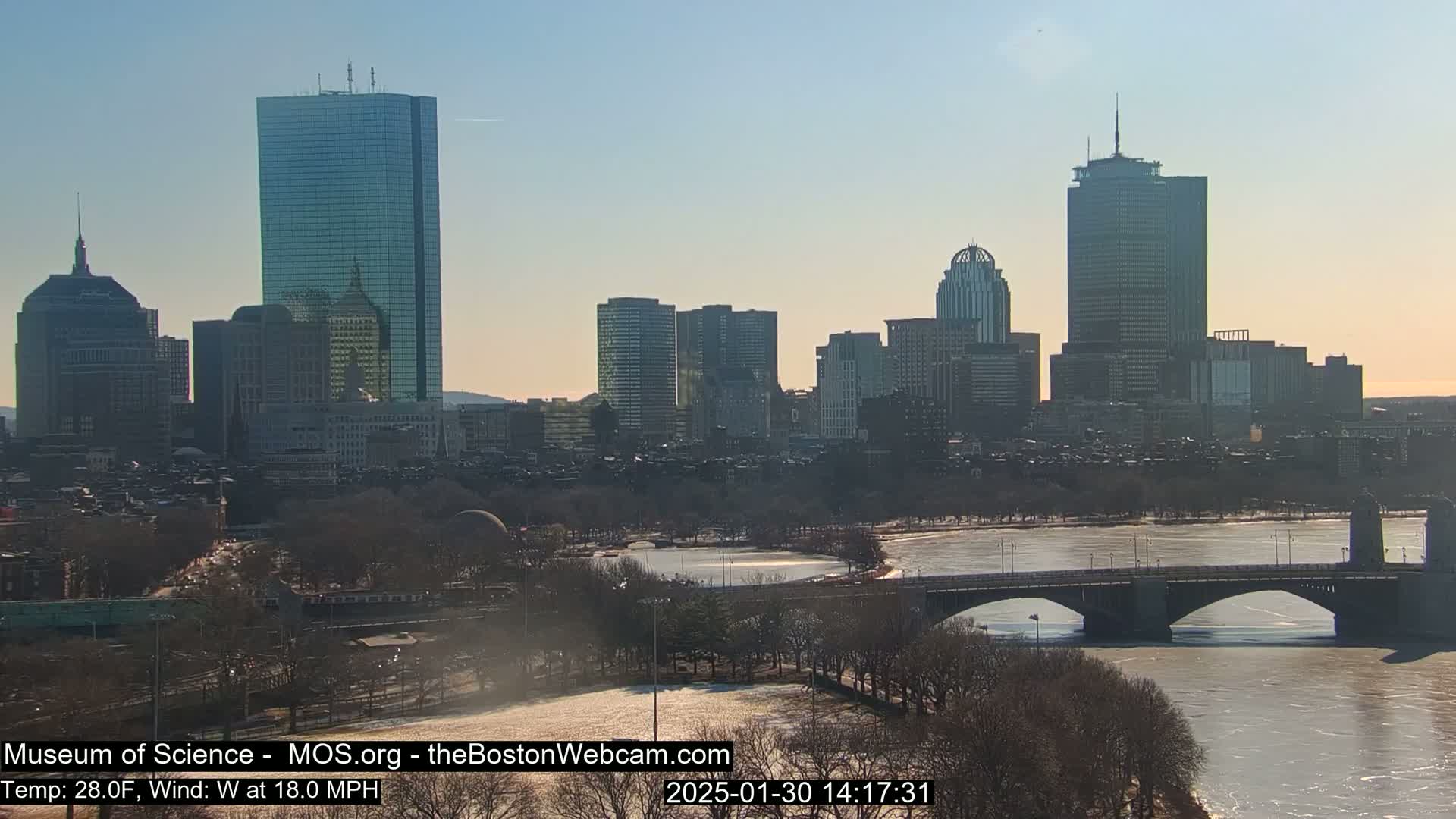 A city skyline with several skyscrapers is seen across a partially frozen river, with a bridge in the foreground under a sunny sky, and a temperature of 28°F and a 18 mph west wind.