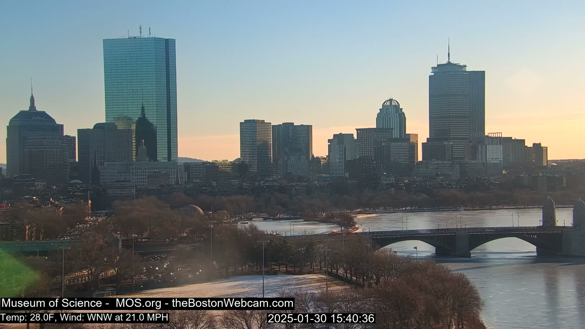 A partly frozen river flows beneath a bridge in front of a cityscape at sunset, with a brisk wind.