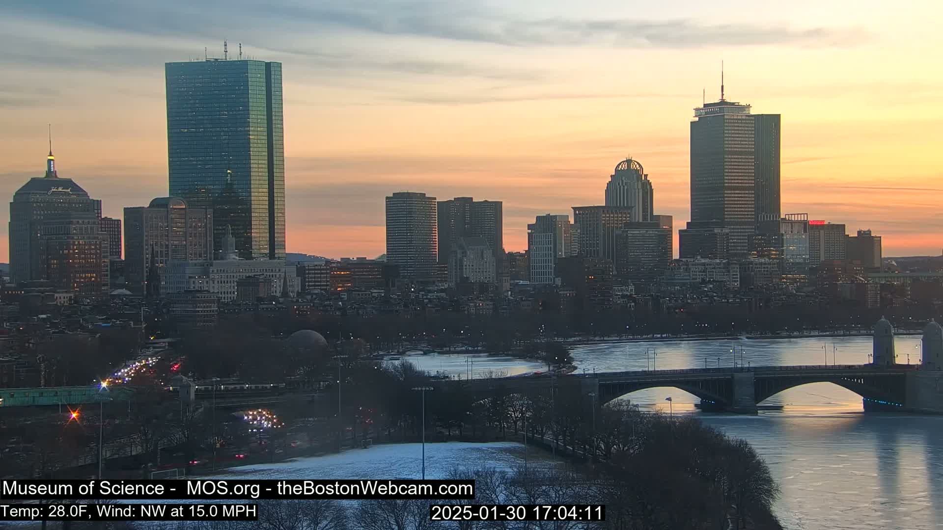 A city skyline at sunset, with a partially frozen river and a bridge in the foreground, under a clear, pale orange and pink sky.