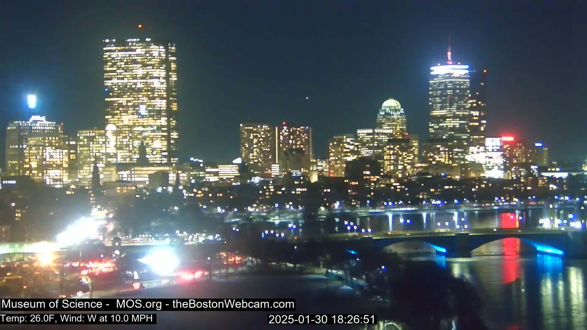 A nighttime view of a city skyline illuminated with lights, across a river with a bridge, under a clear sky.