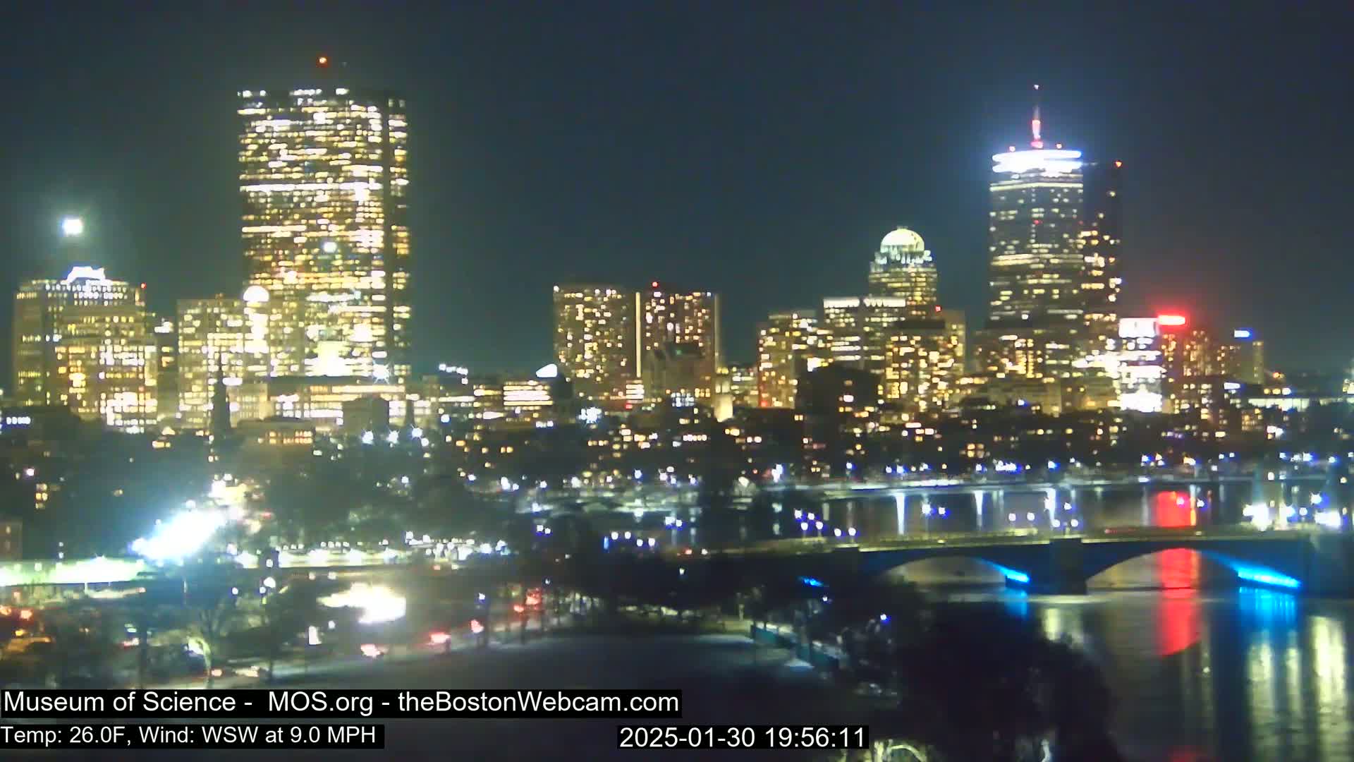 A brightly lit city skyline at night is seen across a river with a bridge, under a clear sky.