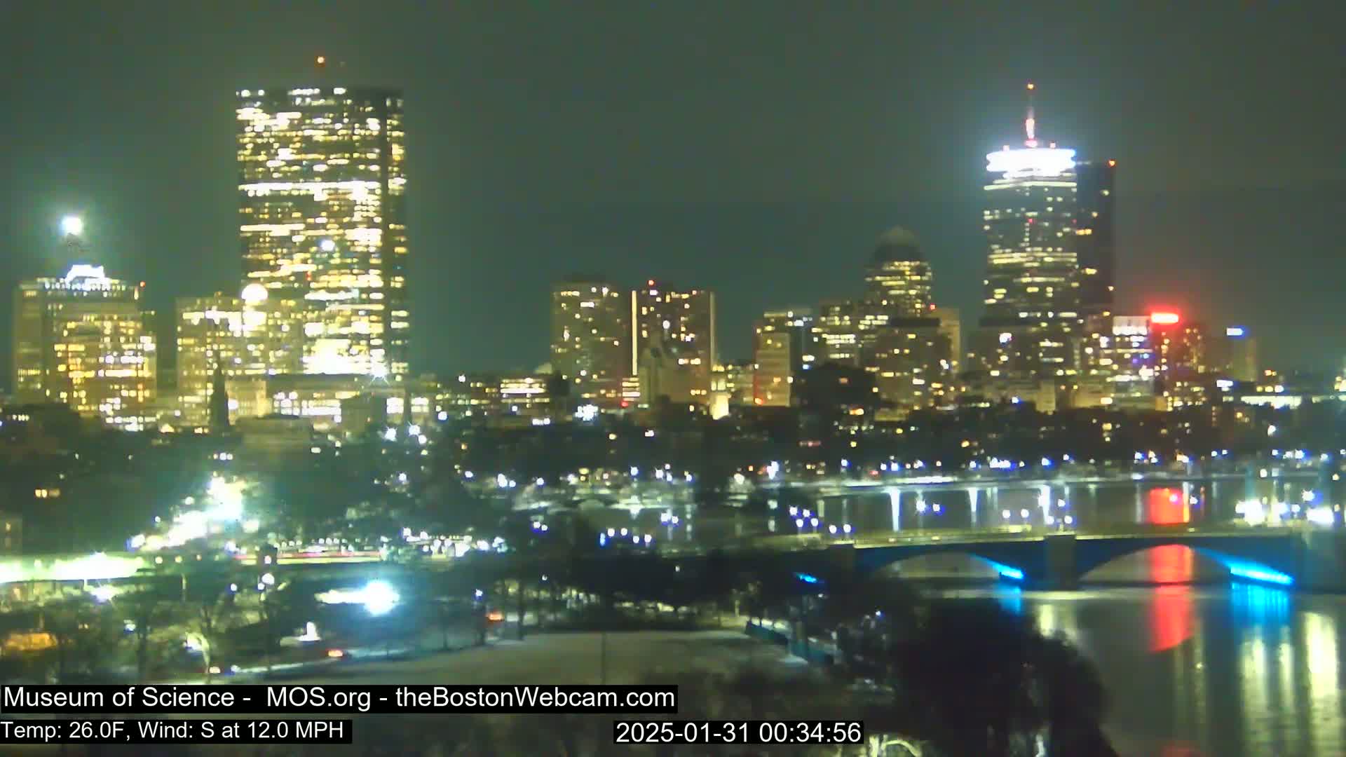A nighttime view of a city skyline with illuminated skyscrapers and bridges across a dark river, under a clear sky with a 26°F temperature and a 12 mph south wind.