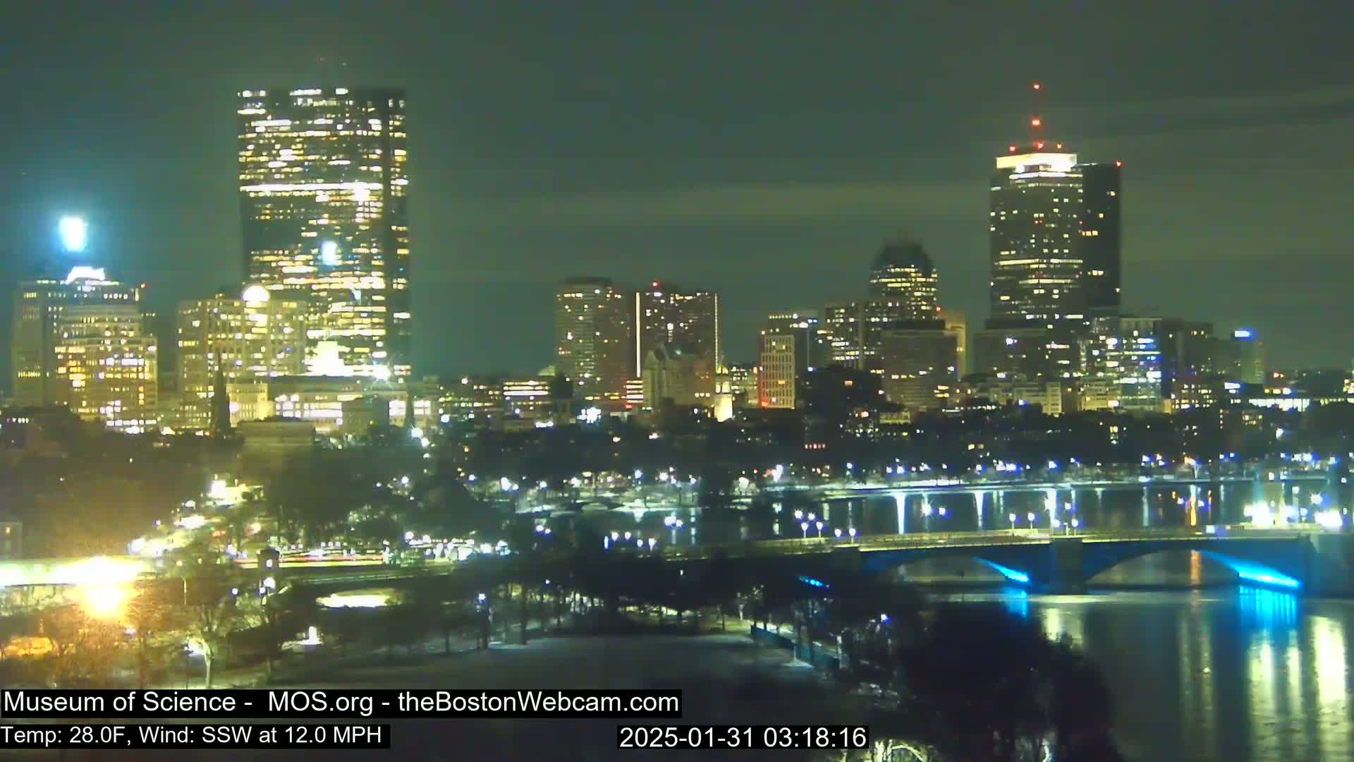 A nighttime cityscape with brightly lit skyscrapers and a bridge illuminated with blue lights, under a clear, dark sky, with a temperature of 28°F and a south-southwest wind.