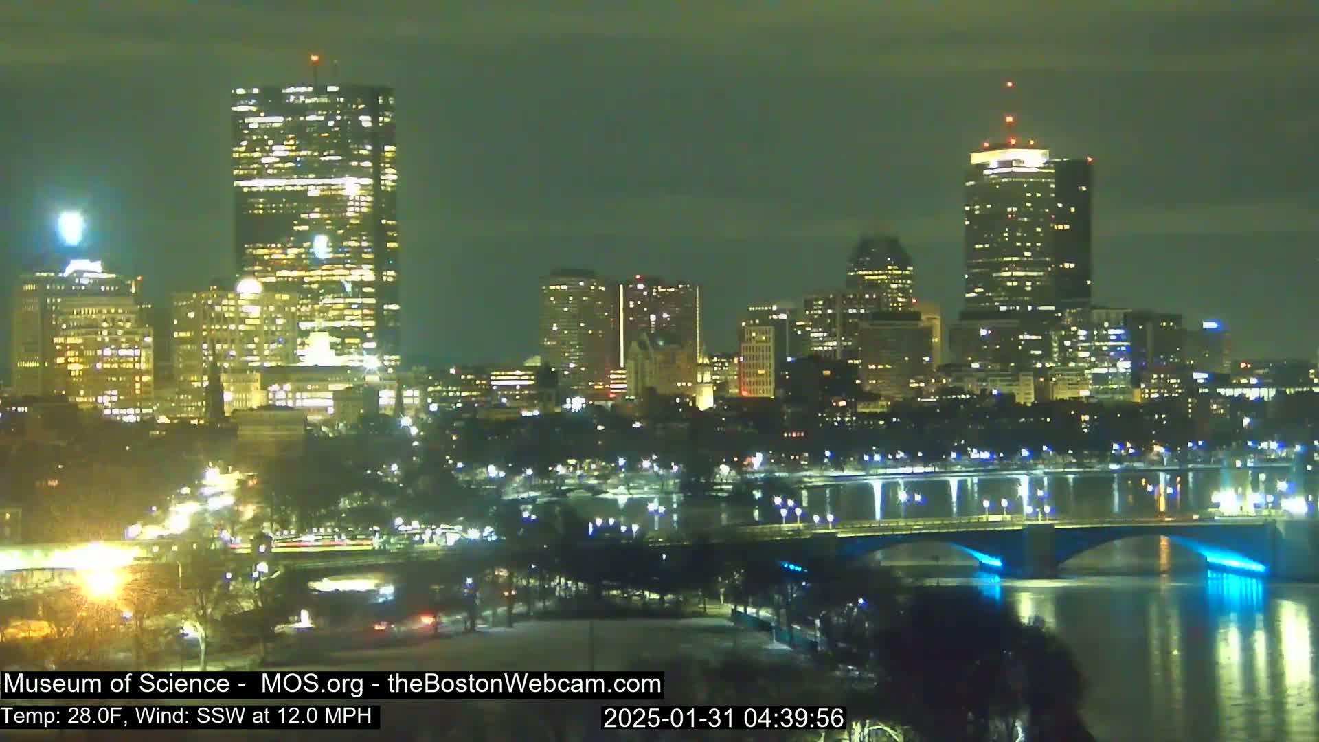 A nighttime cityscape with illuminated skyscrapers and a bridge spanning a body of water under a clear, dark sky, with a temperature of 28°F and a south-southwest wind at 12 mph.
