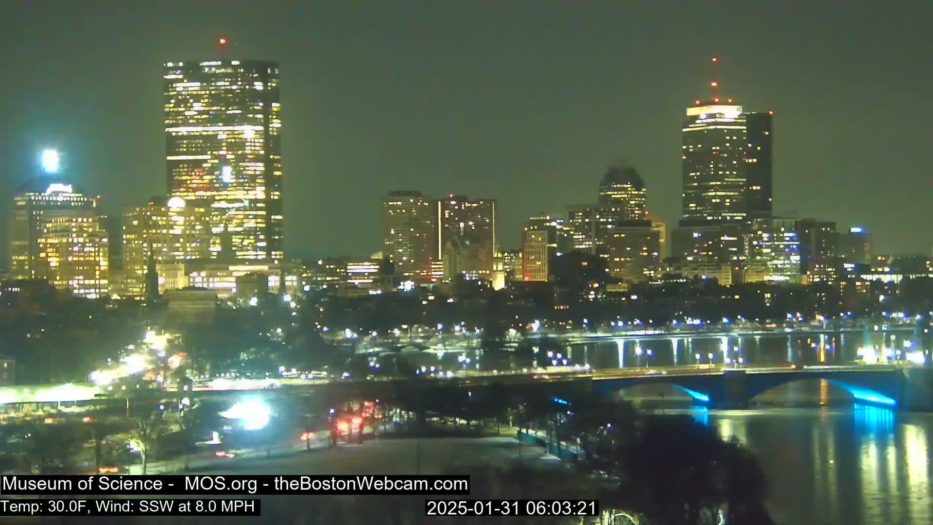 A nighttime view of a city skyline with illuminated skyscrapers and bridges over a river, under a clear, dark sky with a temperature of 30 degrees Fahrenheit and a south-southwest wind.