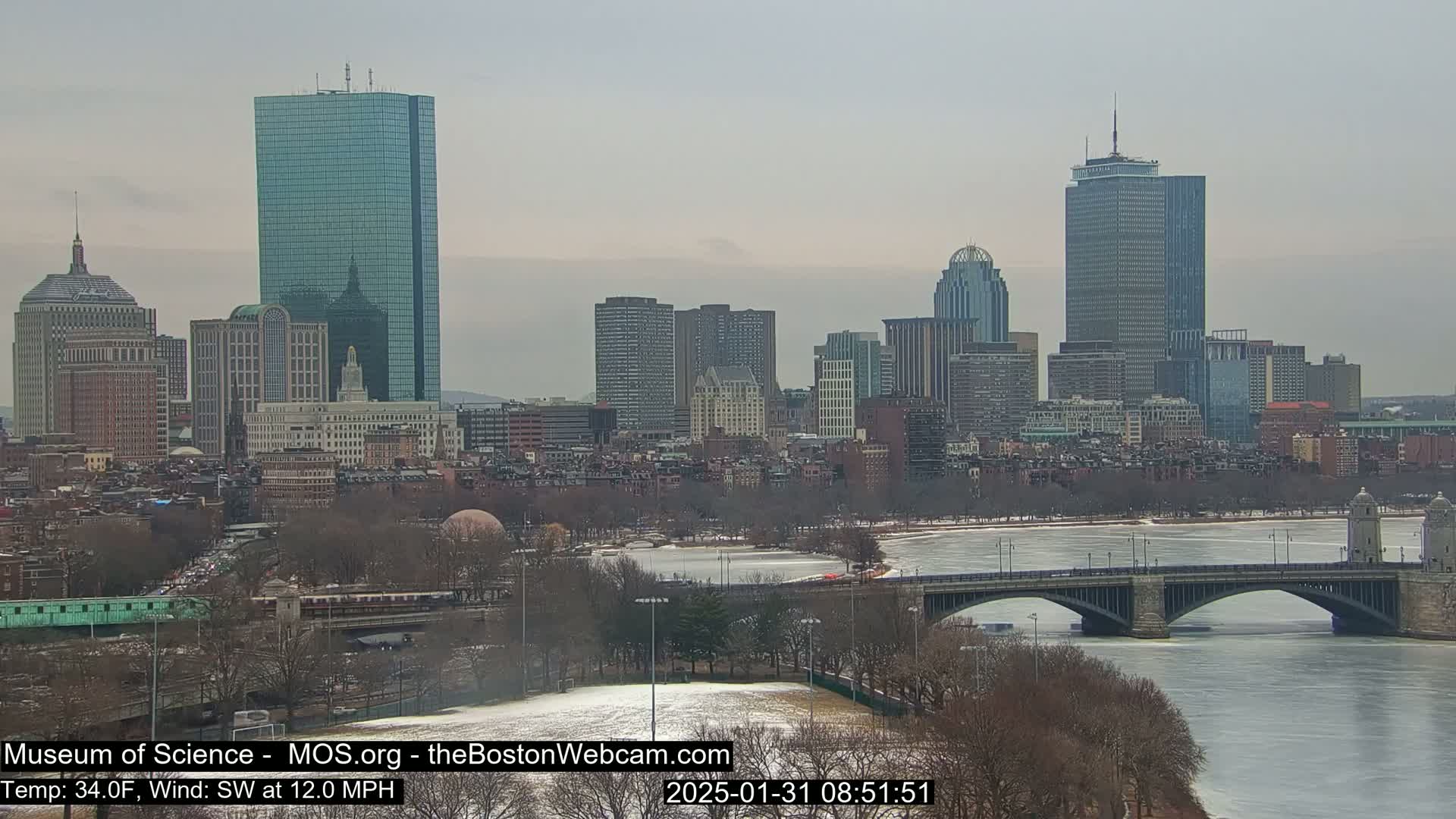 A snow-dusted cityscape with numerous skyscrapers overlooks a partly frozen river and bridge under an overcast sky.