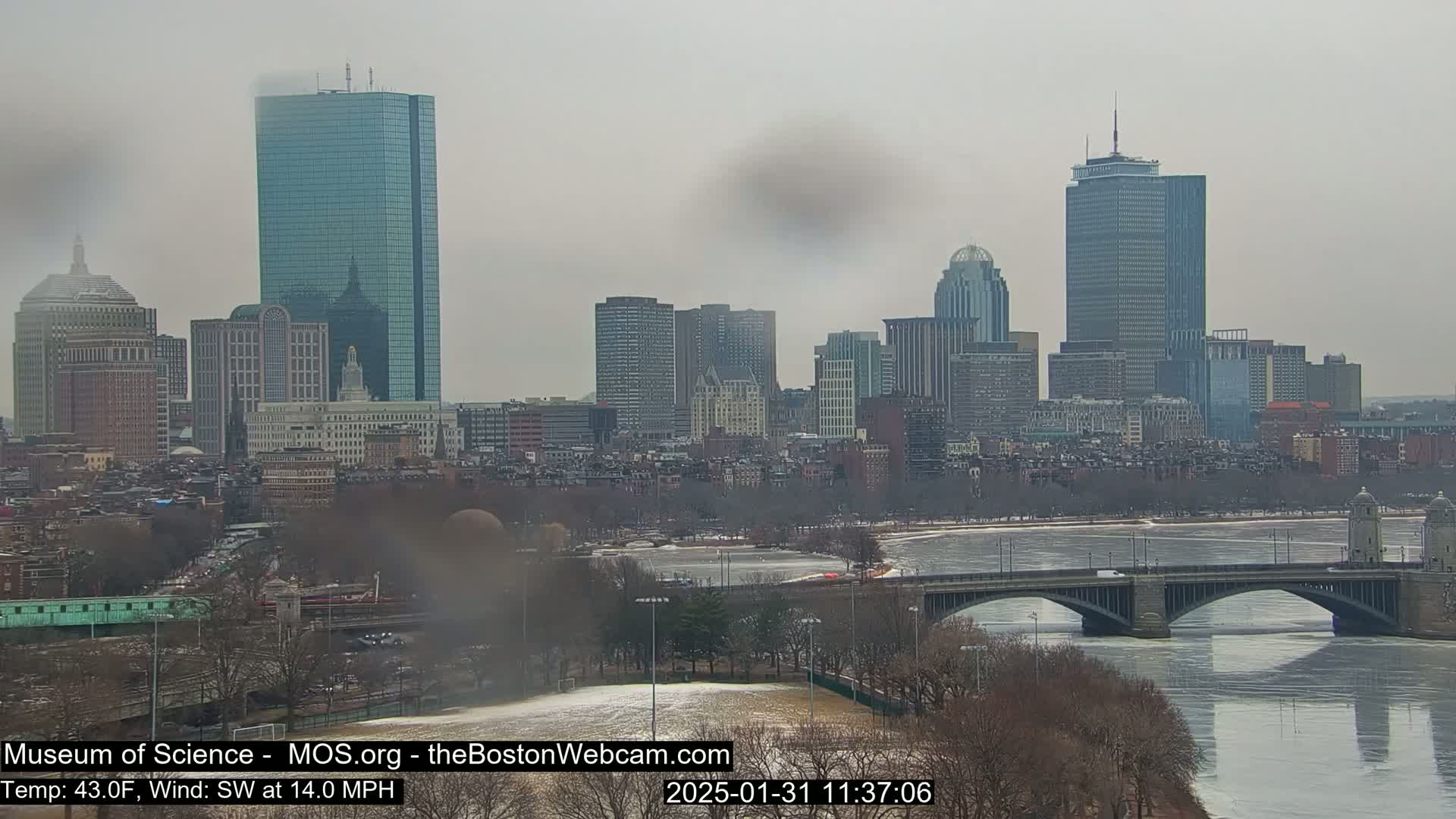 A partly cloudy, cool day shows a cityscape across a river with a bridge, partially frozen, and bare trees lining the shore.